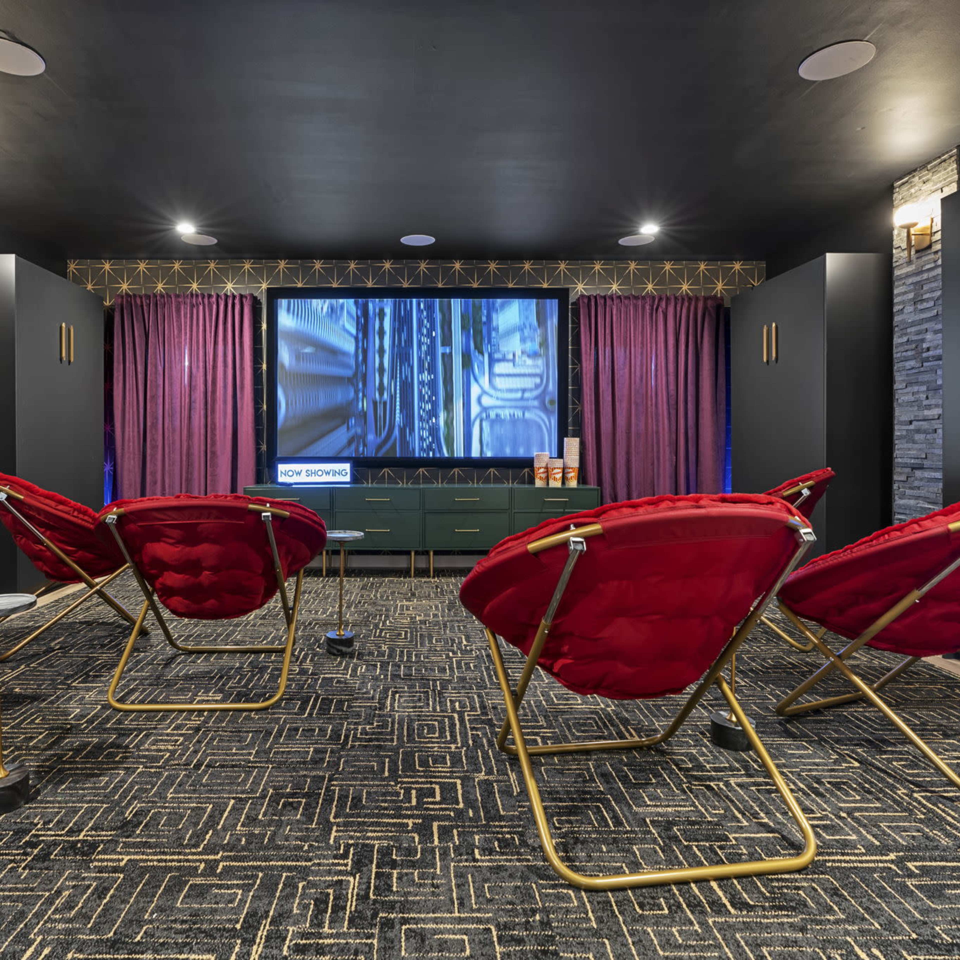 A home theater room features red folding chairs arranged in front of a large screen, with dark walls and patterned carpeting.