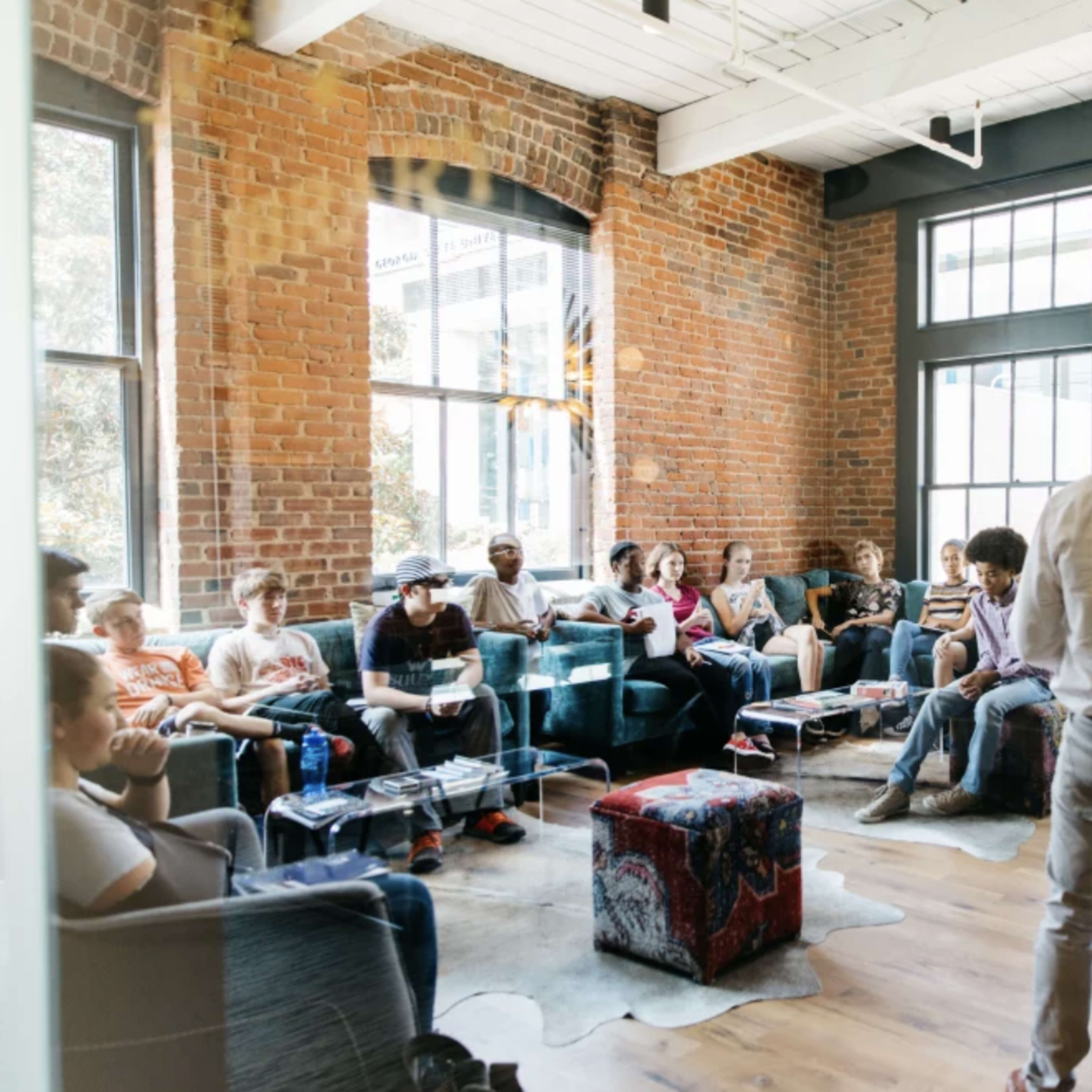 A group of people sits in a bright room with brick walls and large windows, engaged in discussion while a speaker stands in front of them.