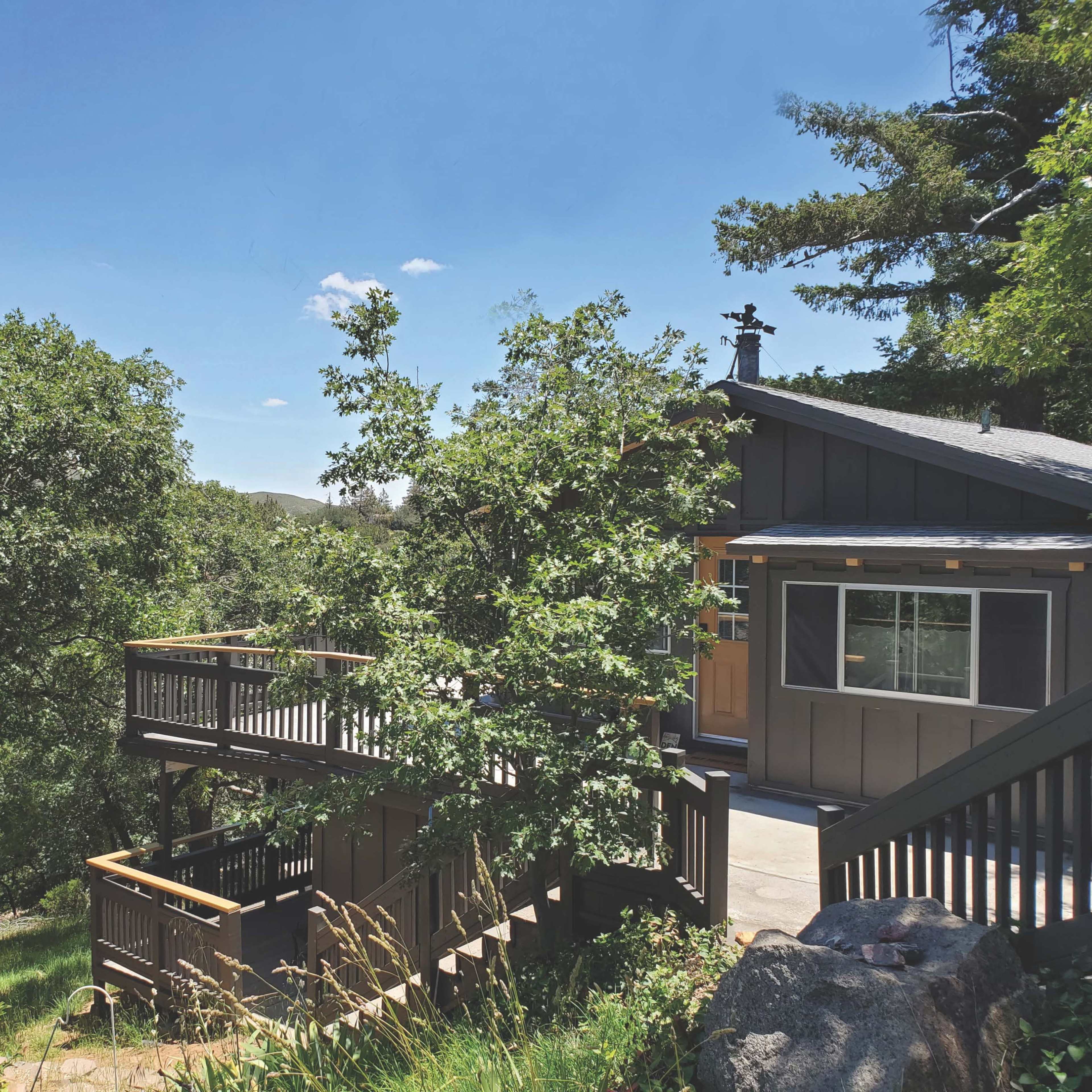 A two-story wooden house is situated among trees on a hillside, featuring a deck that extends from the side of the building.