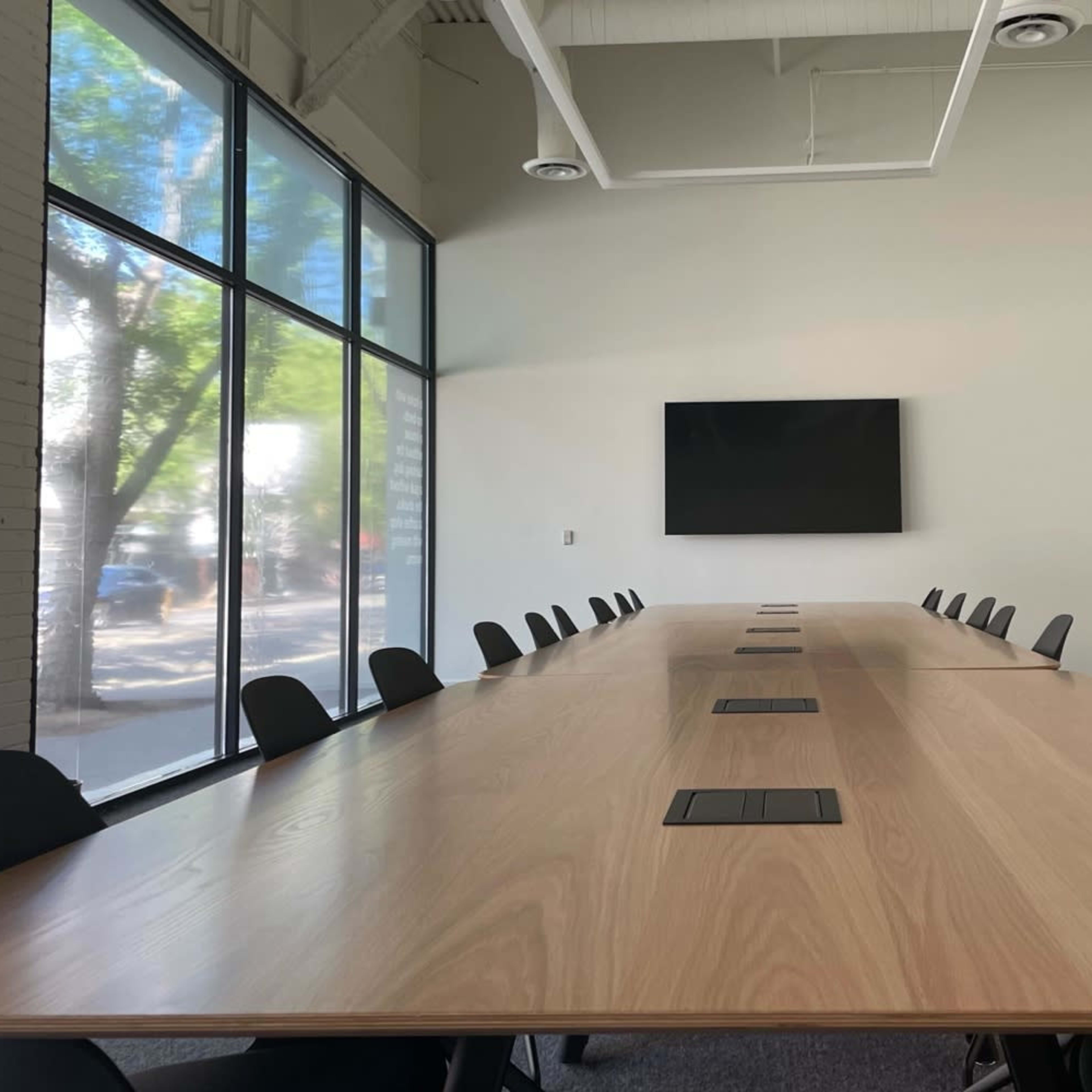 A large, modern conference room features a long wooden table with multiple seats and a wall-mounted screen at the front.