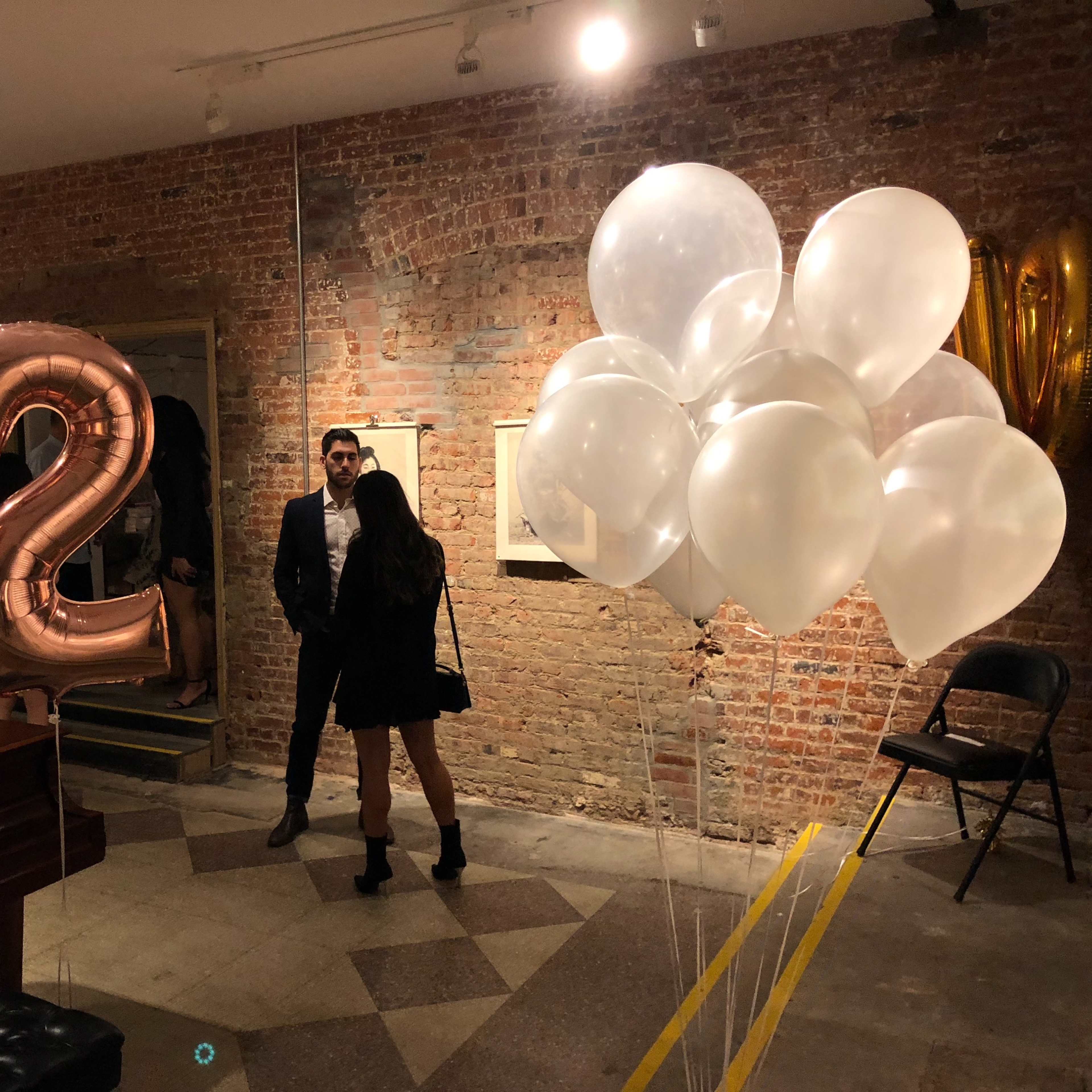 A couple stands conversing in a gallery with large rose gold balloons reading "82" and a cluster of white balloons nearby.