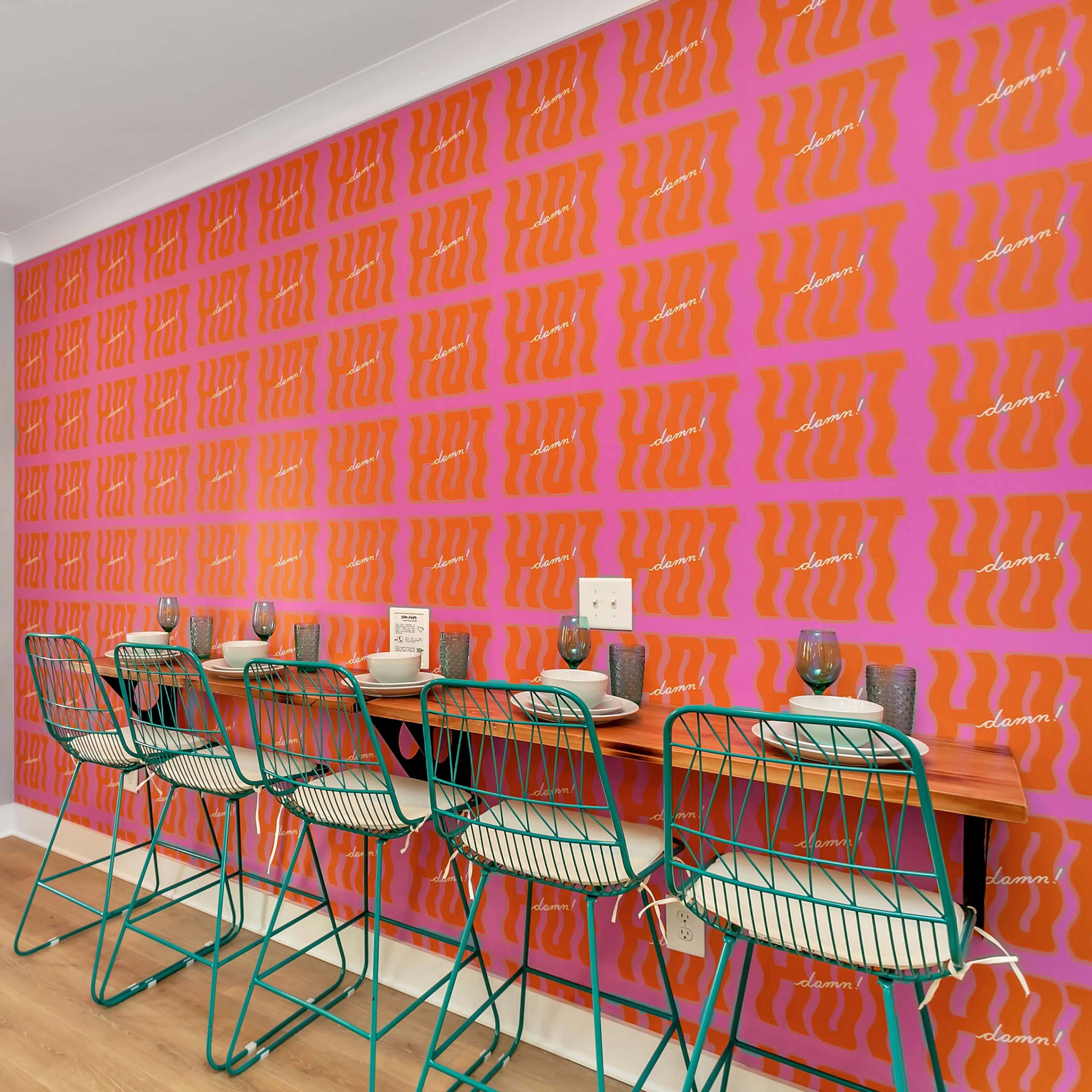 A dining area features a vibrant orange patterned wall with four metal stools lined up at a wooden counter near a white door.