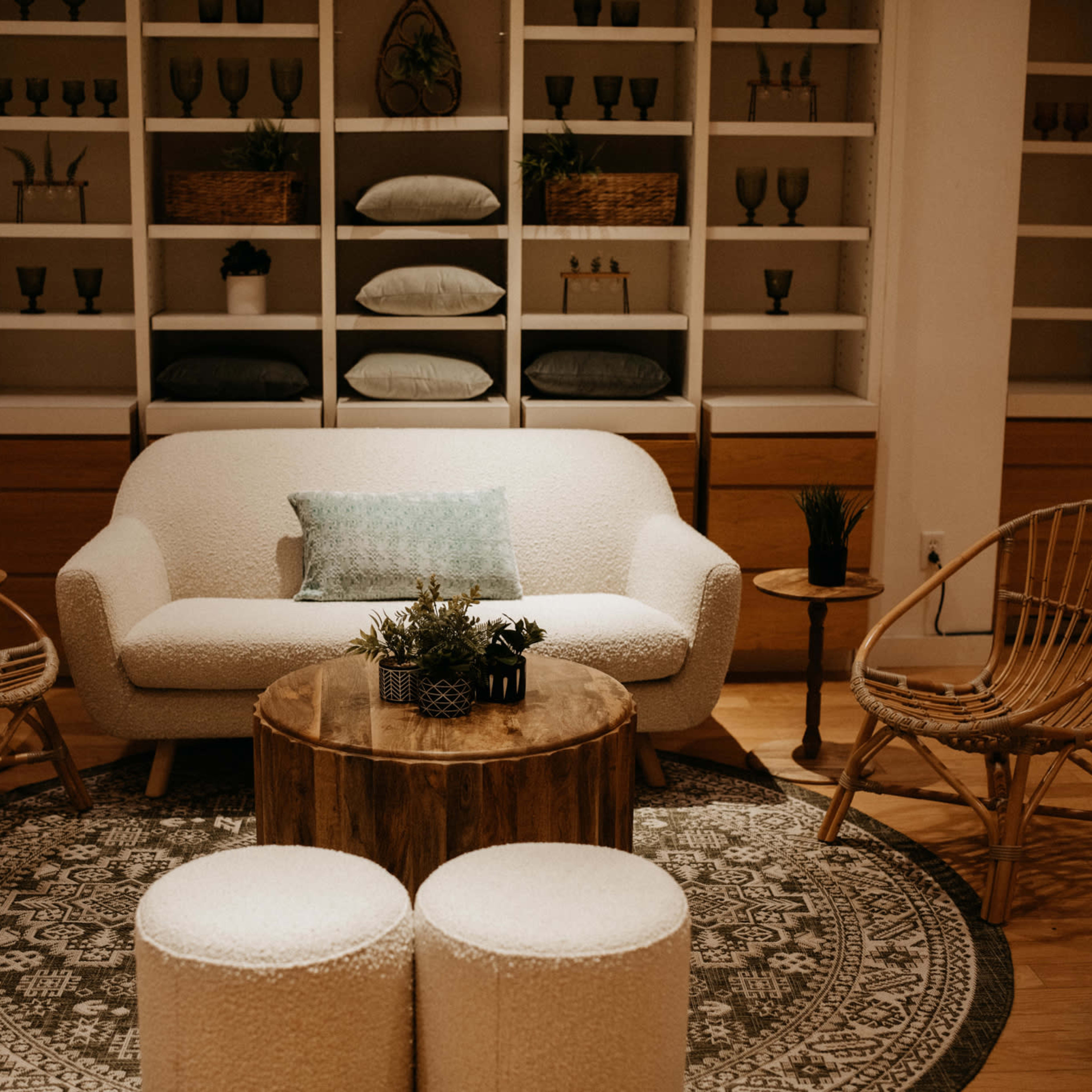 A modern living room features a white sofa, decorative plants, a wooden coffee table, and woven chairs arranged around a round rug.