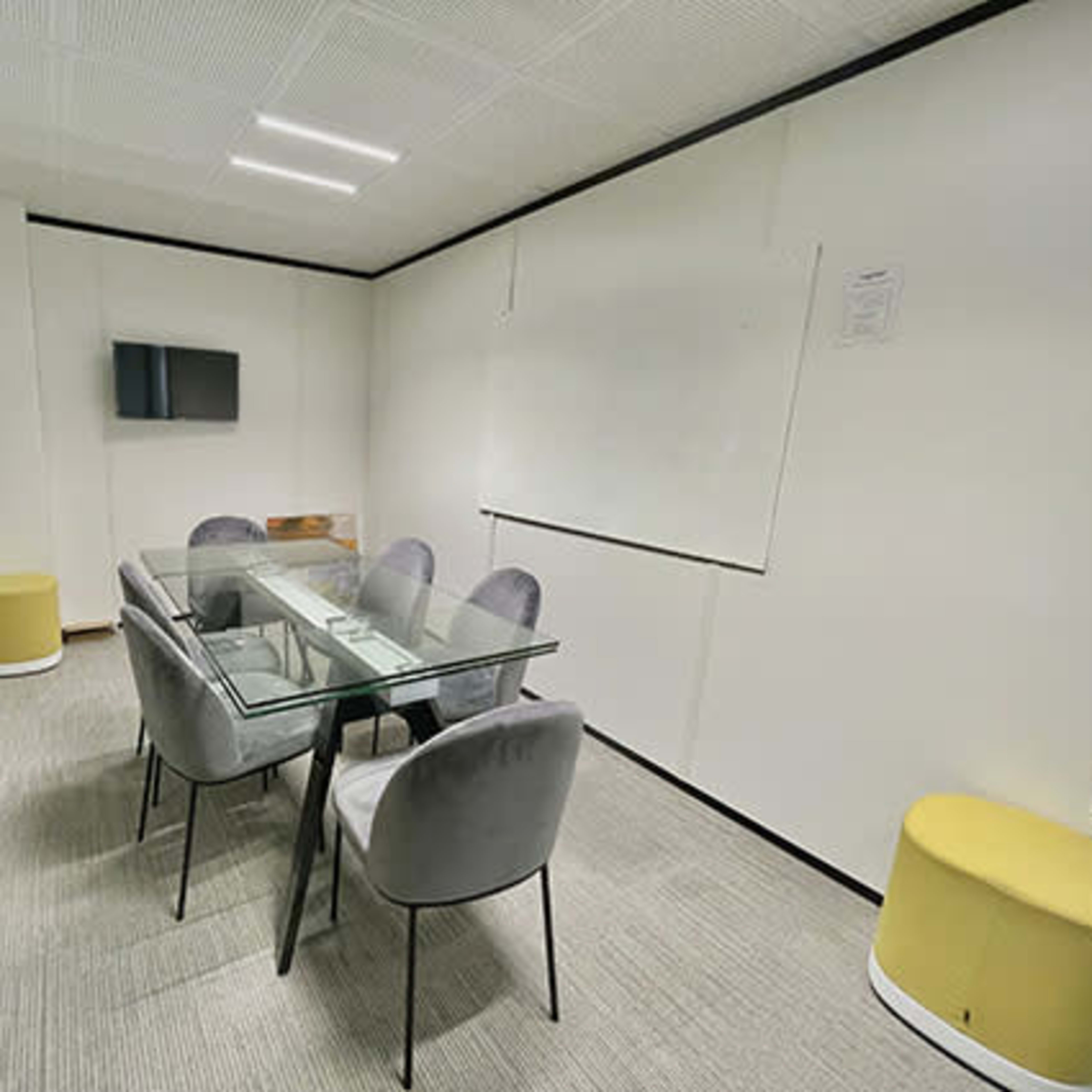 A conference room features a glass table surrounded by gray chairs, with a television and a whiteboard on the walls.