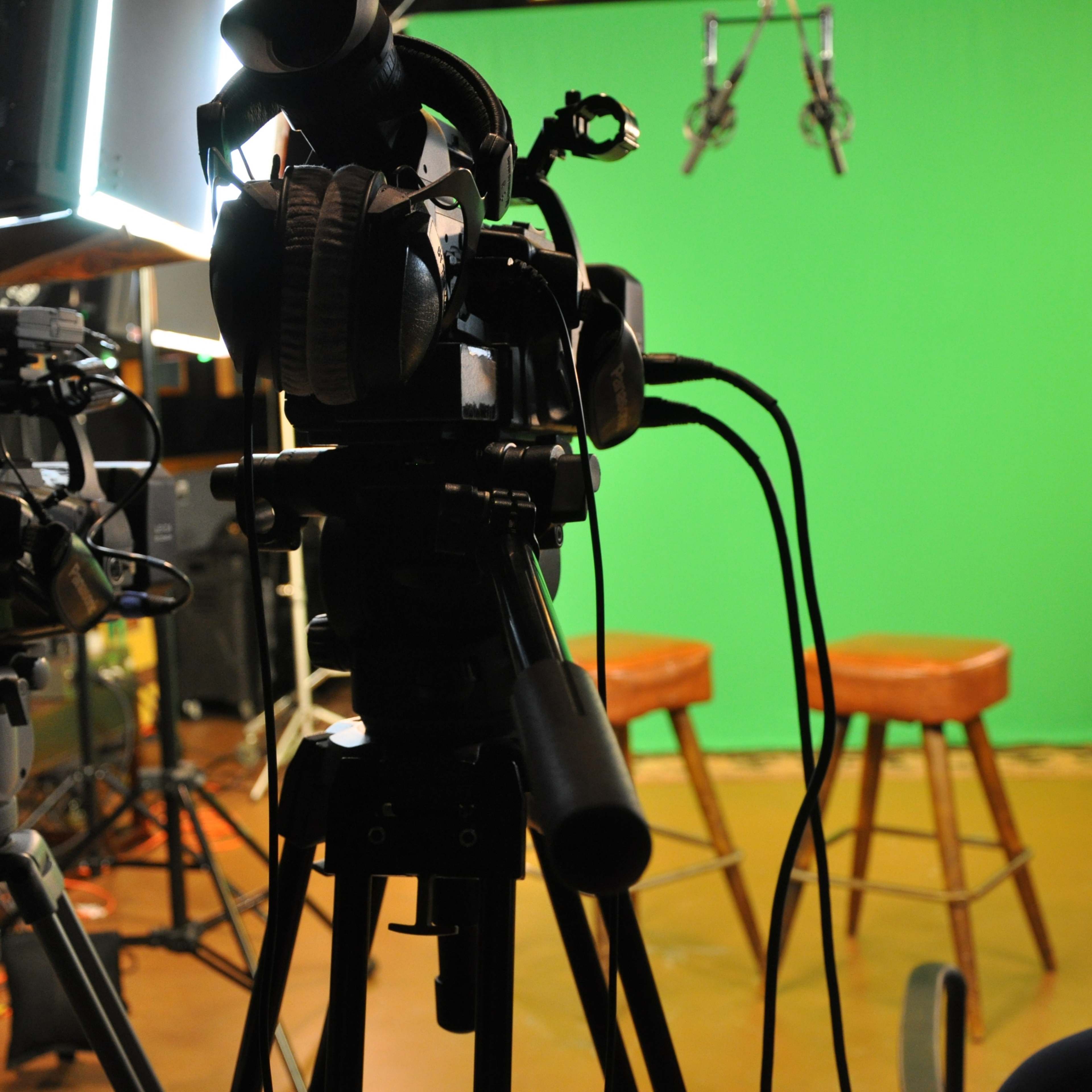 A camera setup is positioned in front of two empty stools against a green screen backdrop in a studio setting.