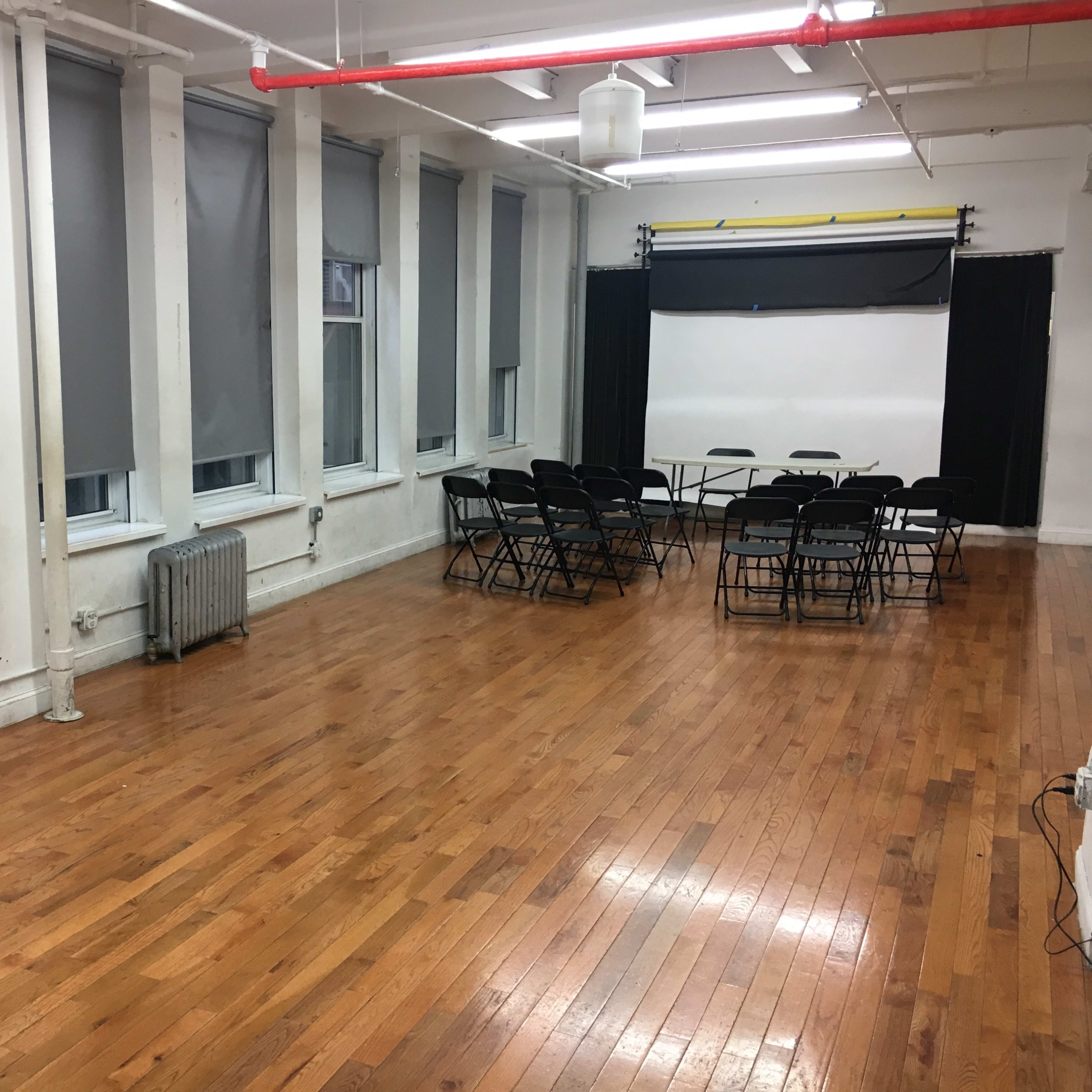 A large, empty room with hardwood floors, gray blinds, and a setup of black chairs arranged around a table at the back.