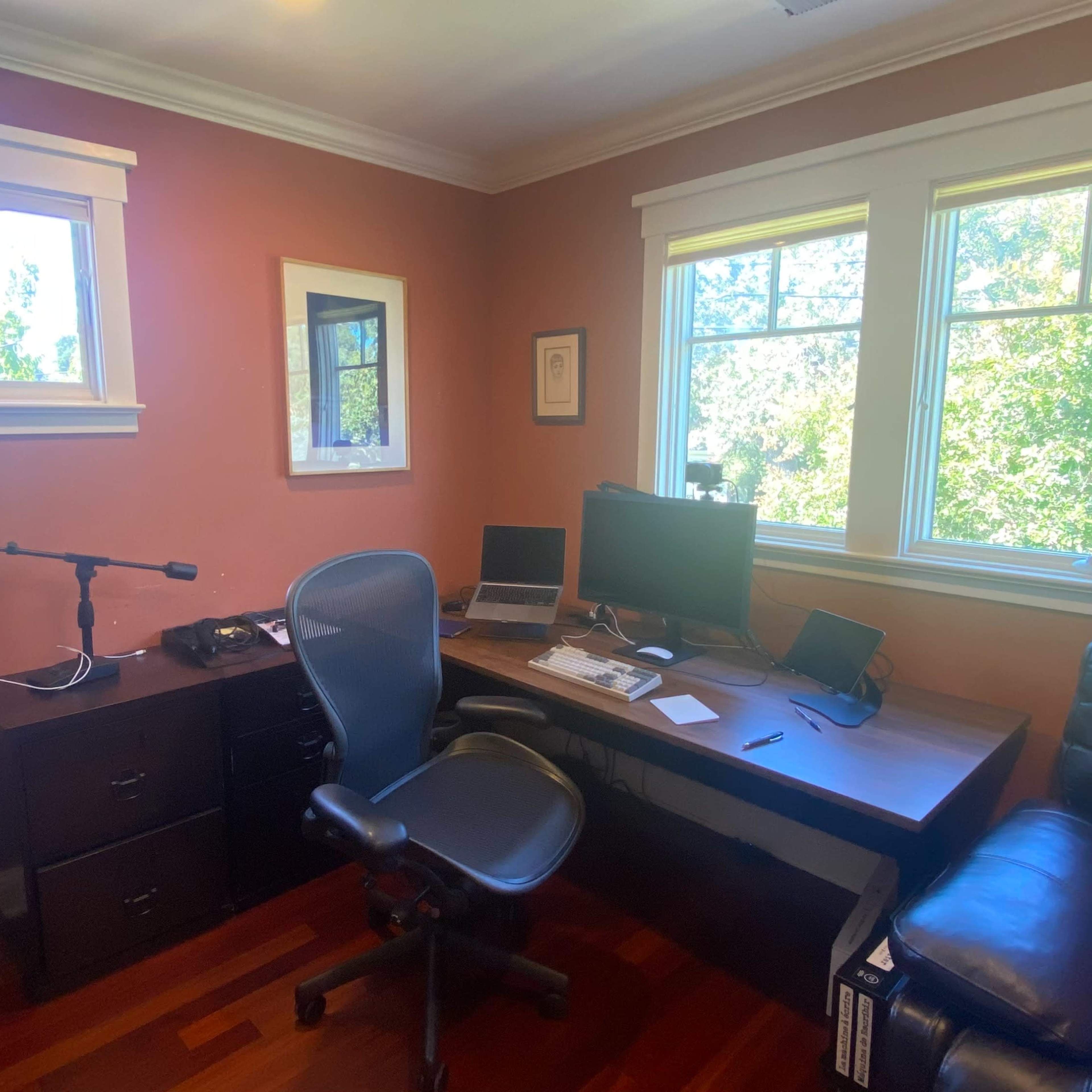 A home office features a desk with two computer monitors, a microphone, and a seating chair, along with large windows allowing natural light to enter the room.