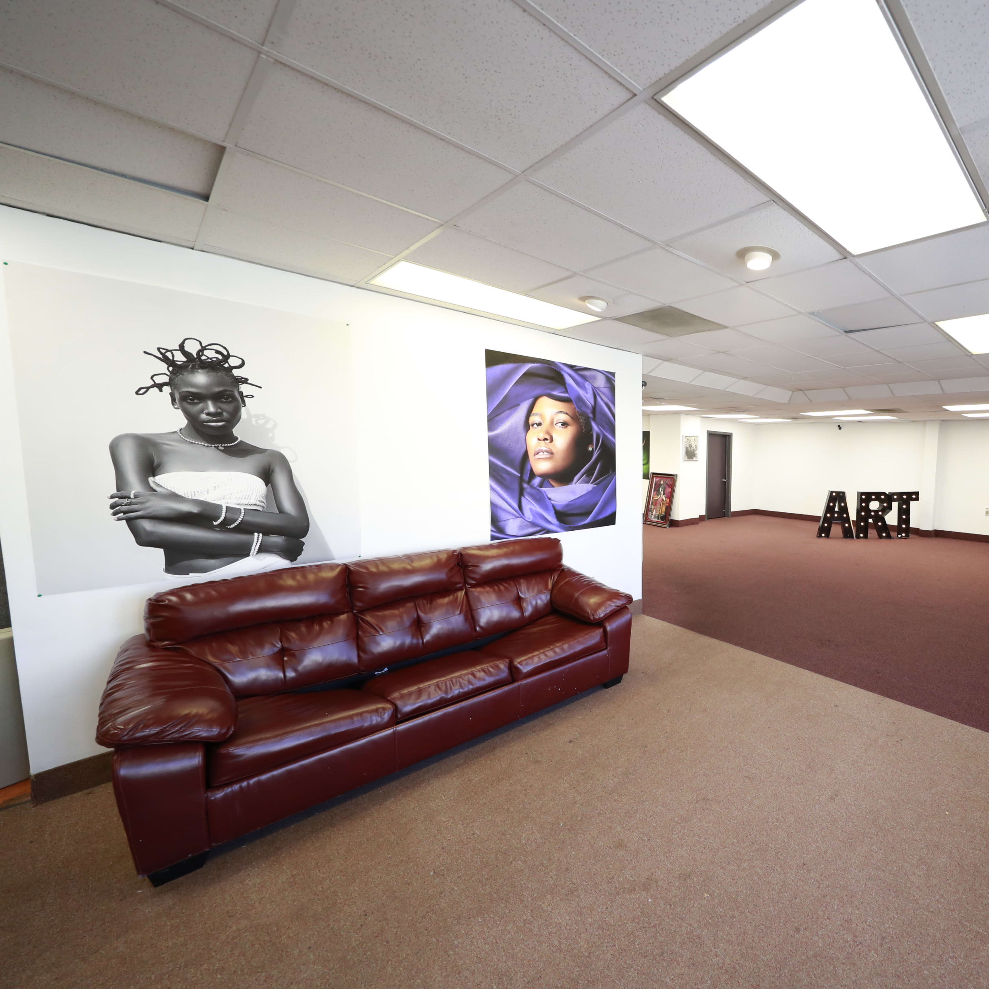 A spacious interior room with a brown leather sofa, large wall posters of women, and the word "ART" made of wooden letters on the floor.