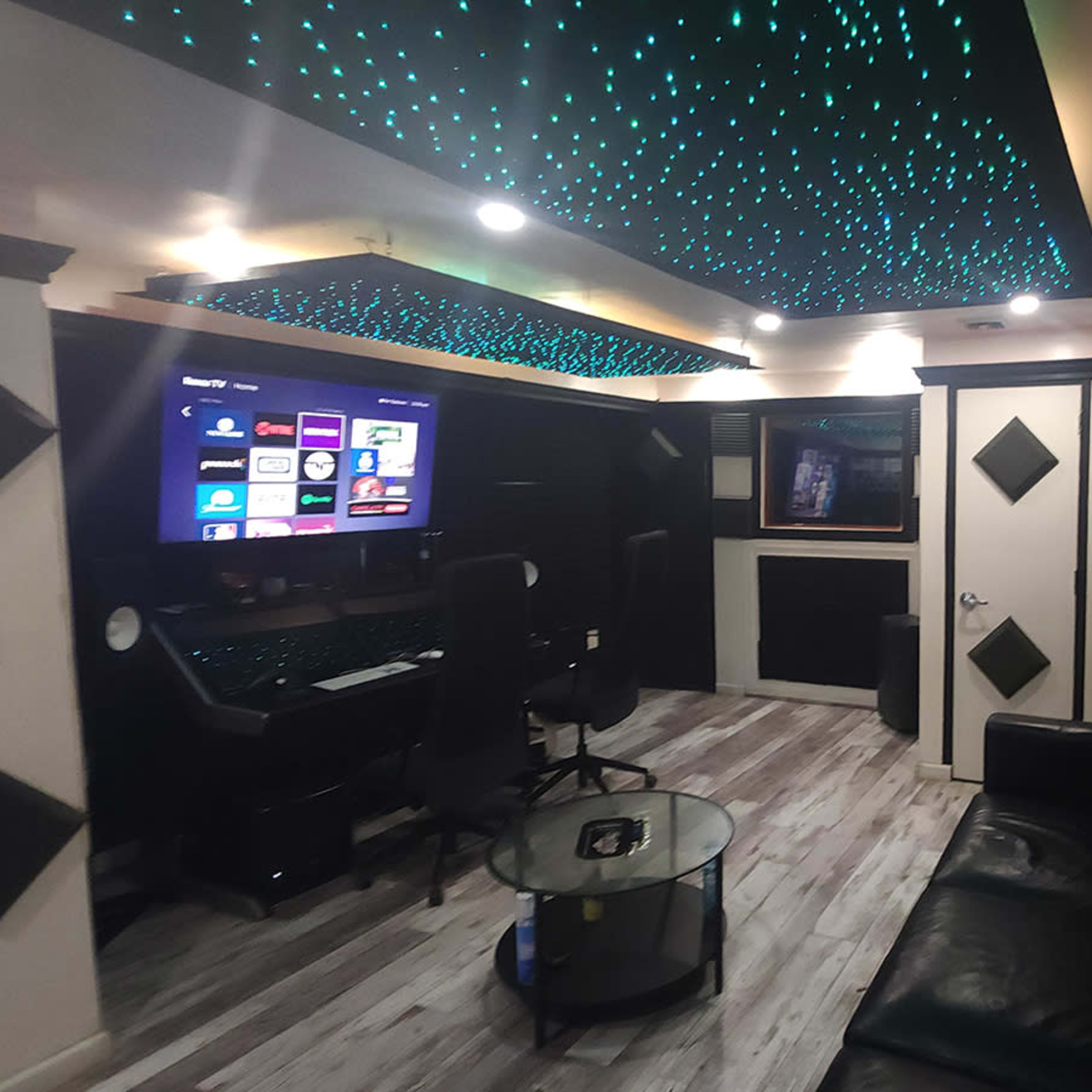 A modern recording studio with a black leather sofa, a glass coffee table, and a workstation equipped with a computer and large screen, illuminated by a starry ceiling.