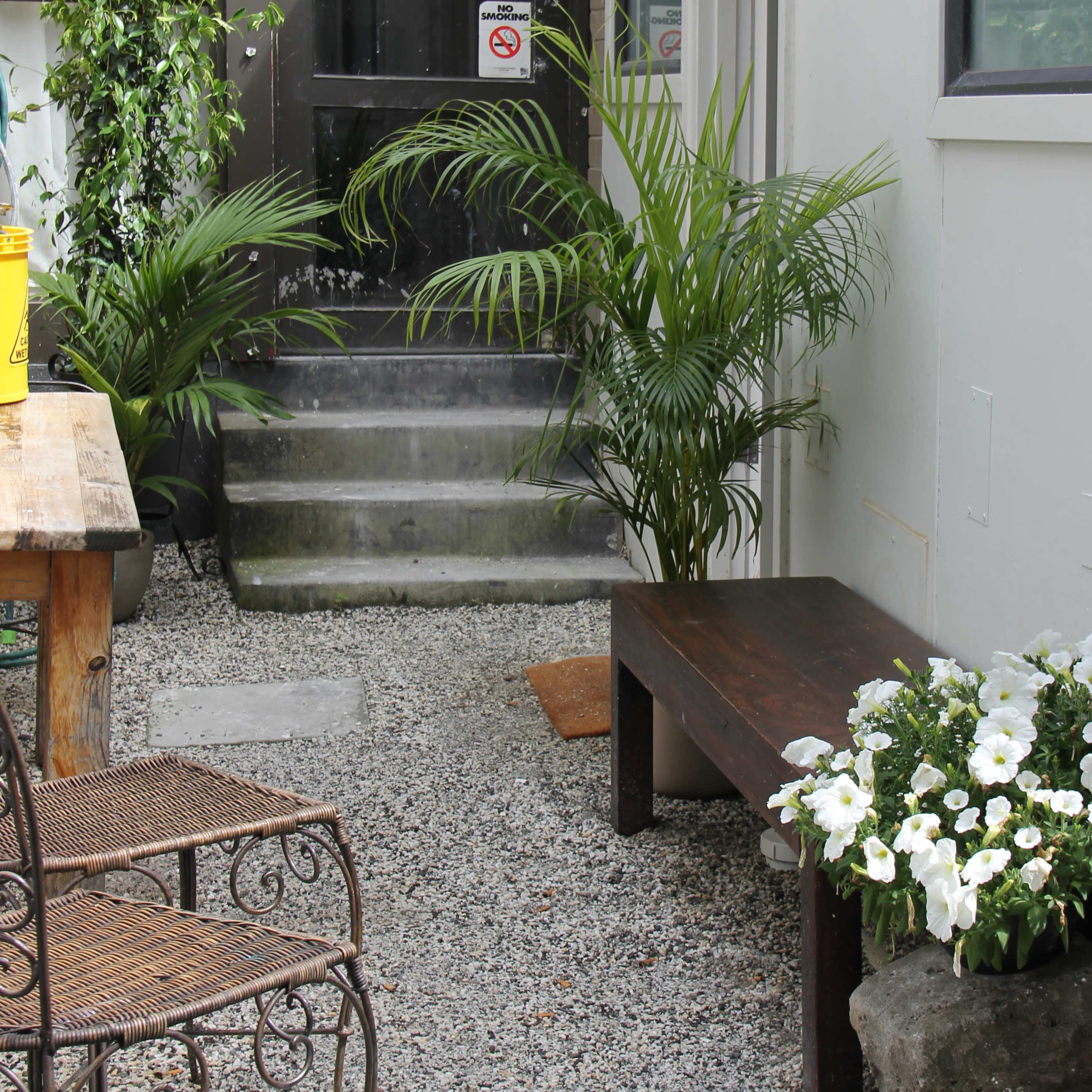 A narrow outdoor pathway with a wooden bench, a potted plant, and white flowers beside a staircase leading to a door.