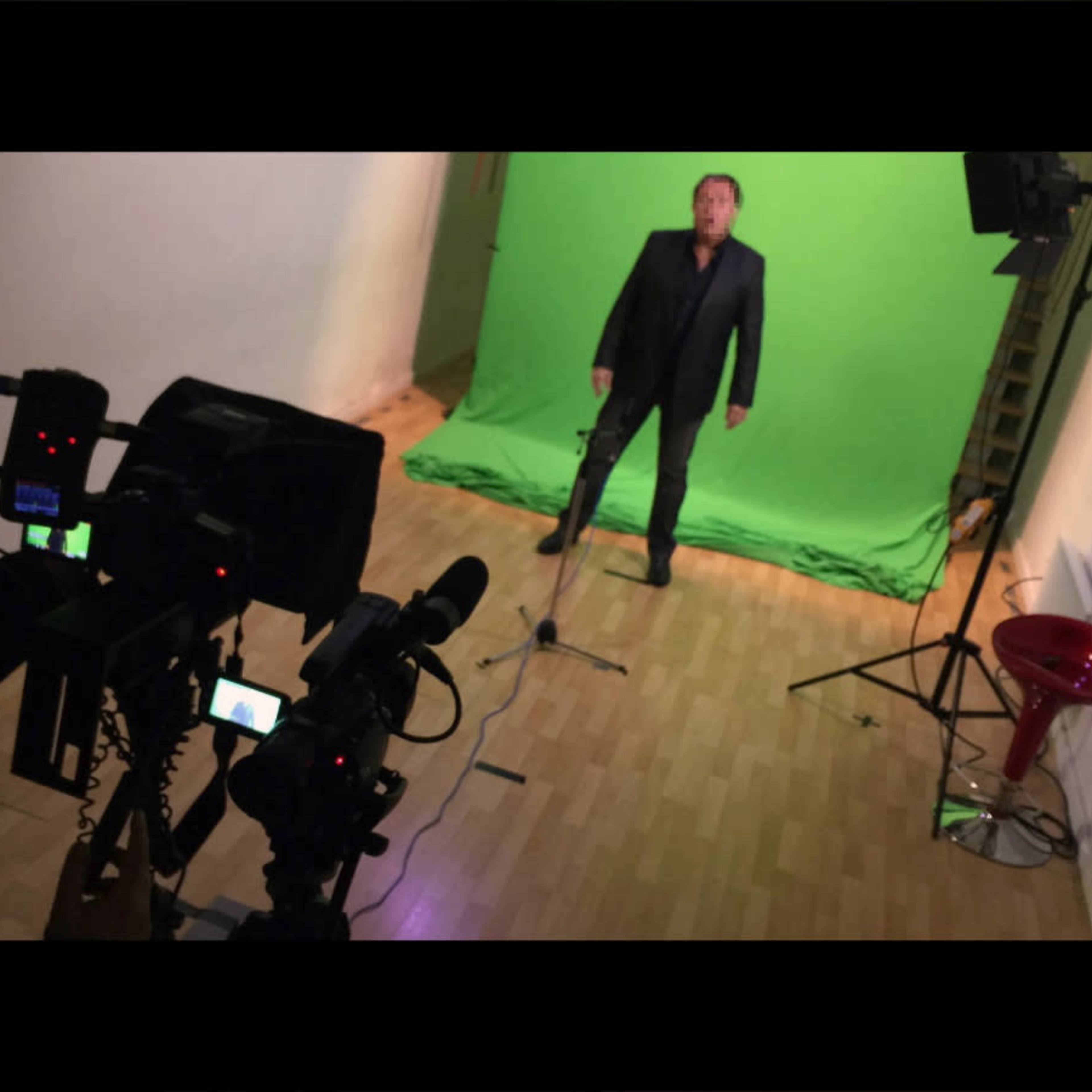A man stands in front of a green screen in a studio, with cameras and a microphone positioned nearby.