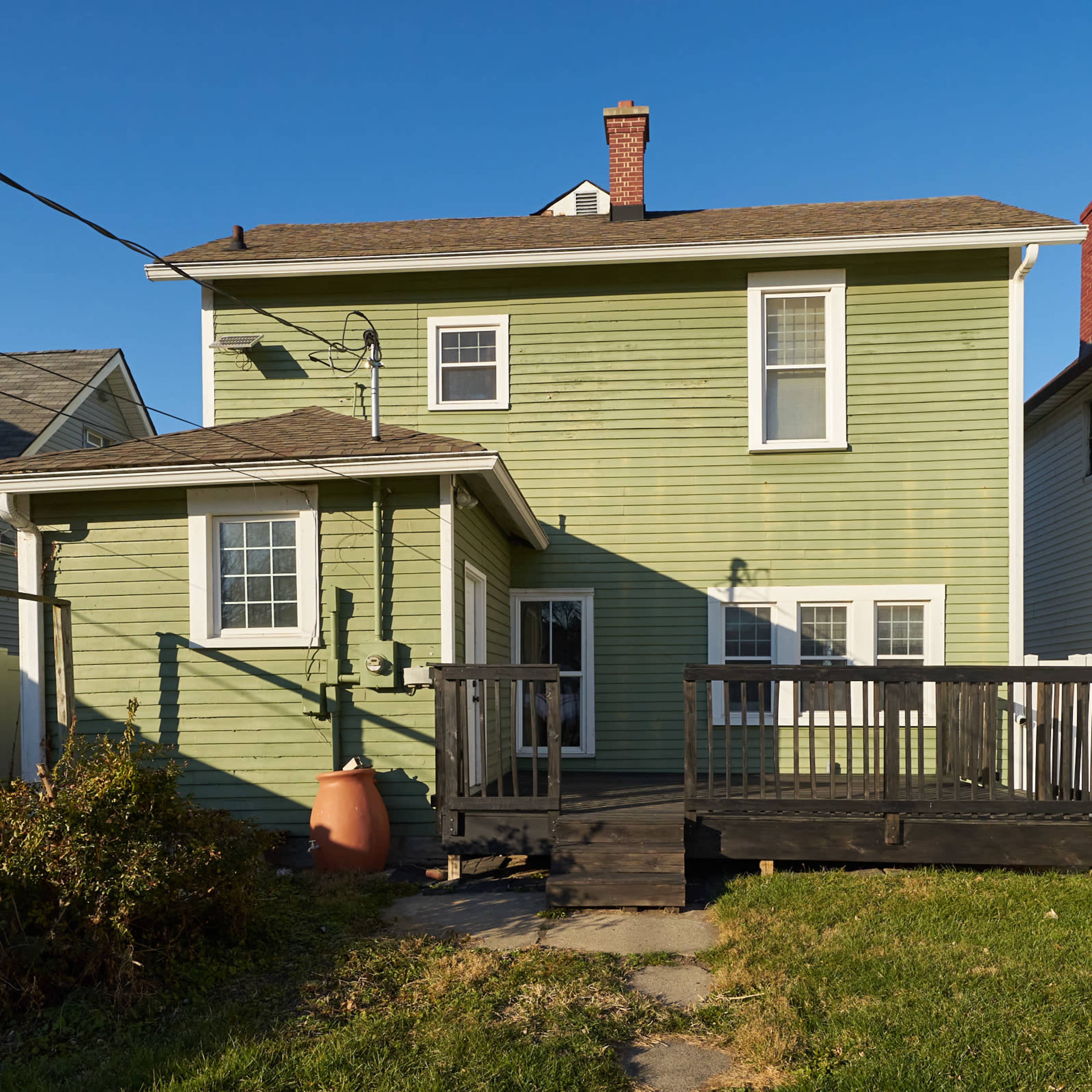 A two-story green house with a wooden deck and a fenced yard surrounded by neighboring houses.