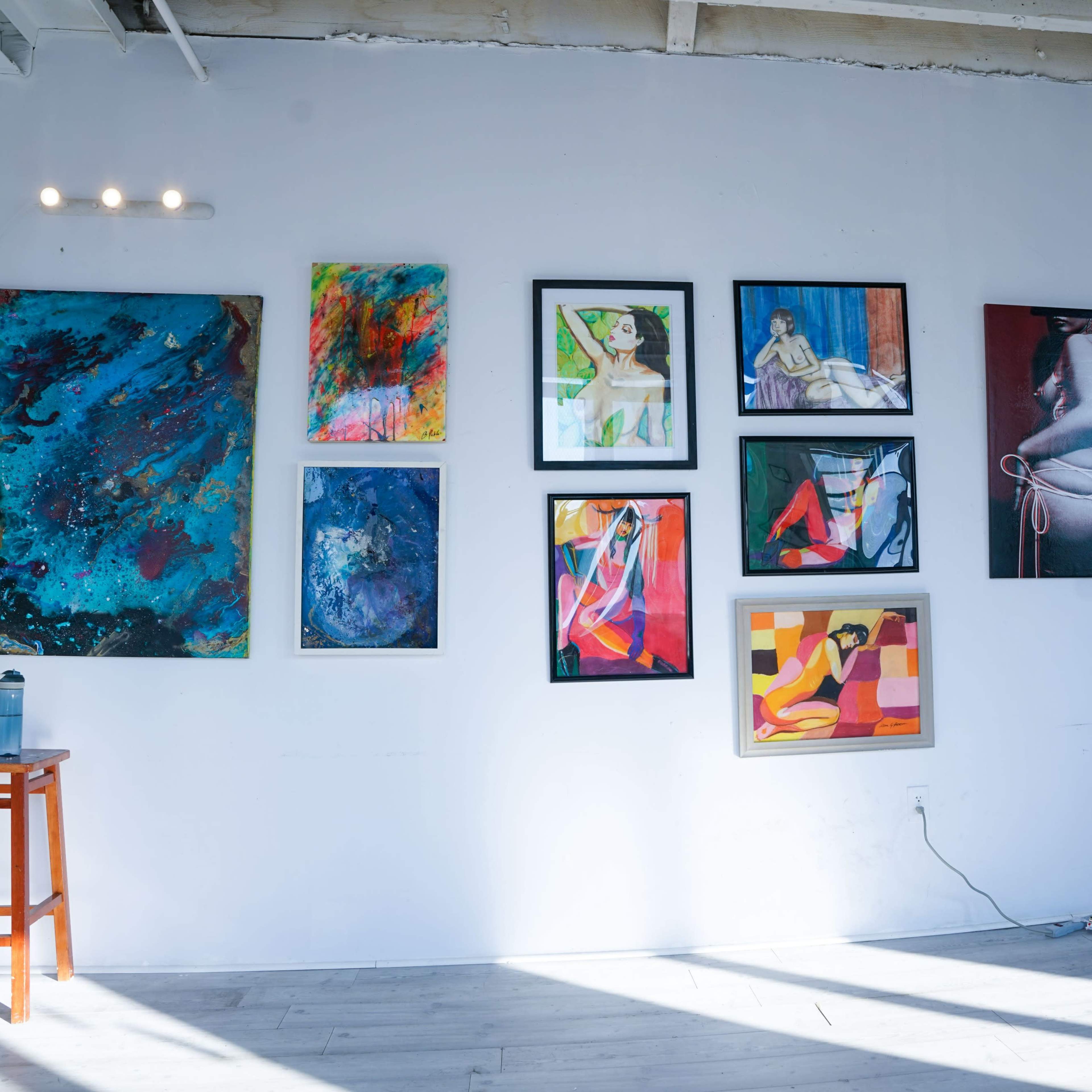 A gallery wall adorned with various framed artworks, including vibrant paintings and drawings, beside a wooden stool and a mirror on the floor.