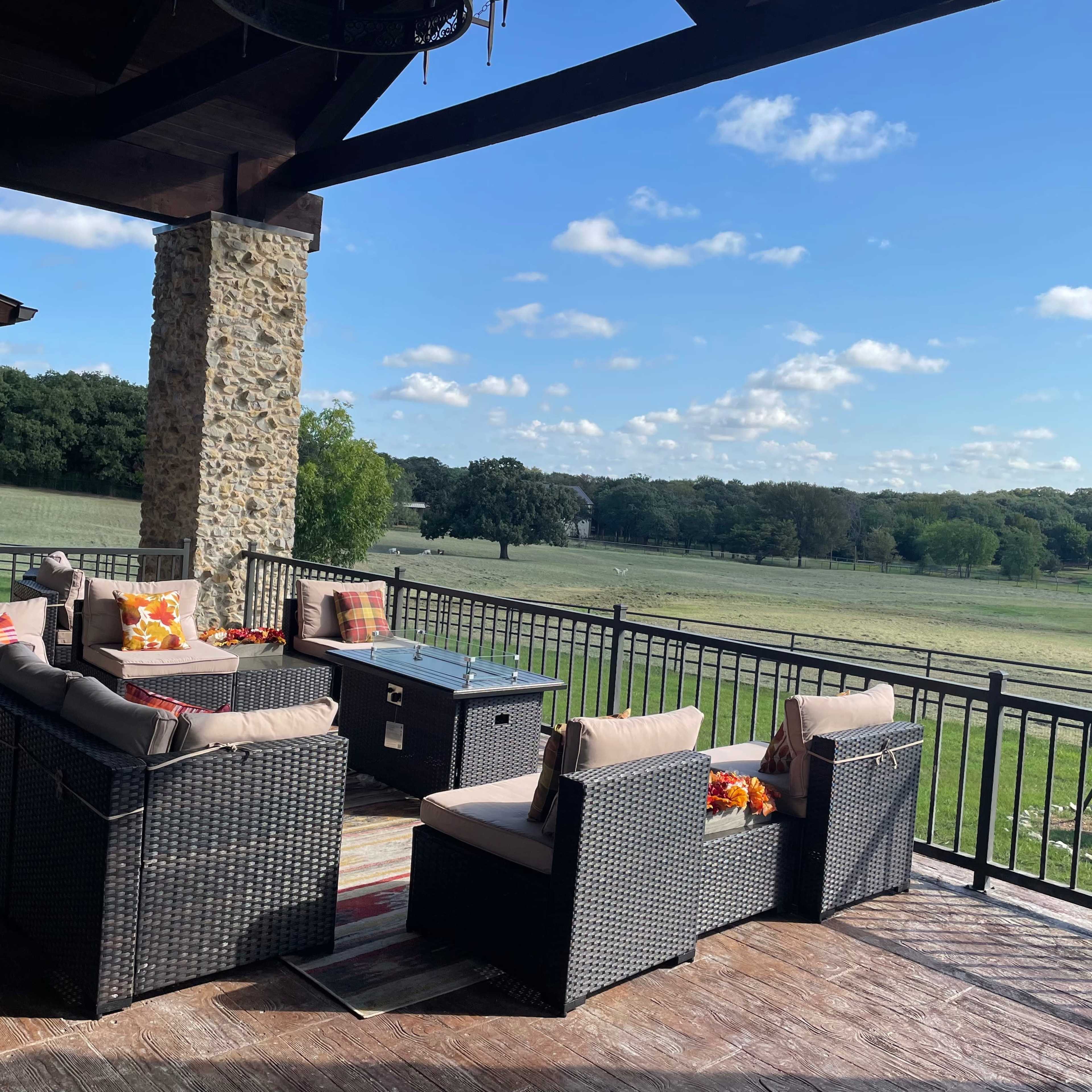A spacious patio features wicker furniture, a coffee table, and a view of a grassy landscape with trees under a partly cloudy sky.