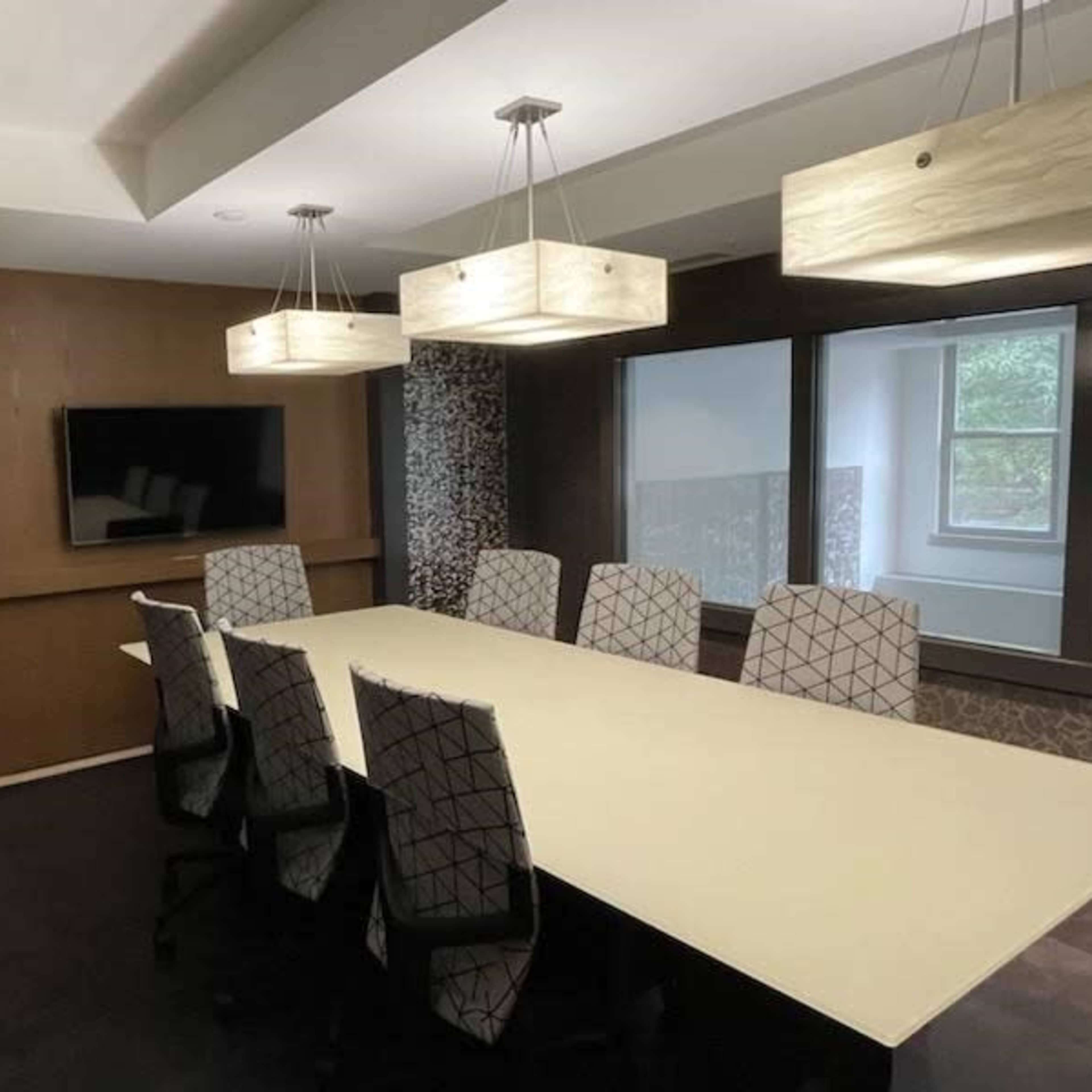 A modern conference room with a long table, eight patterned chairs, a wall-mounted television, and pendant lighting.