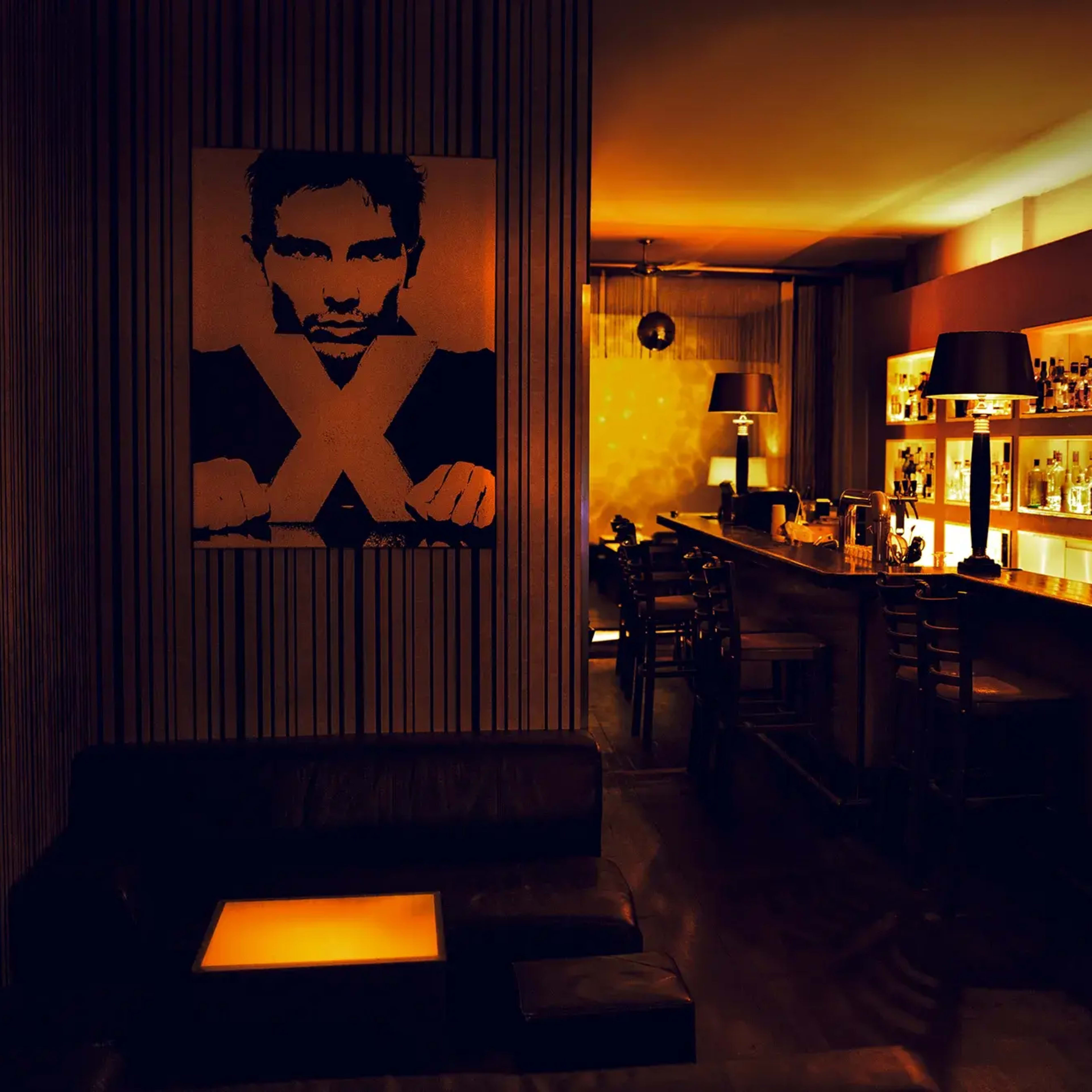 A dimly lit bar features a prominent artwork of a man on the wall, with shelves filled with bottles behind the counter.