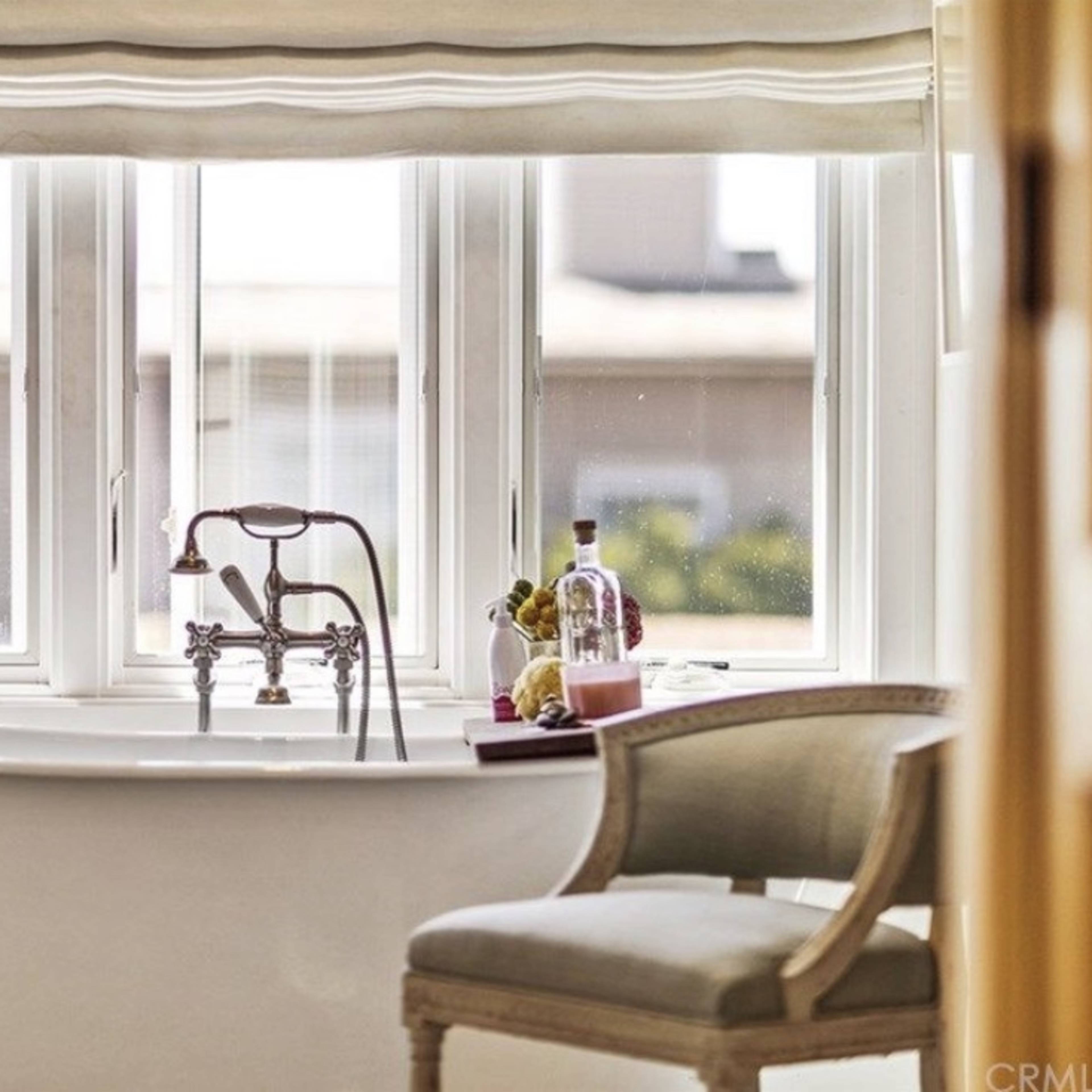 A freestanding bathtub is positioned near large windows, with a potted plant and a chair visible in the scene.