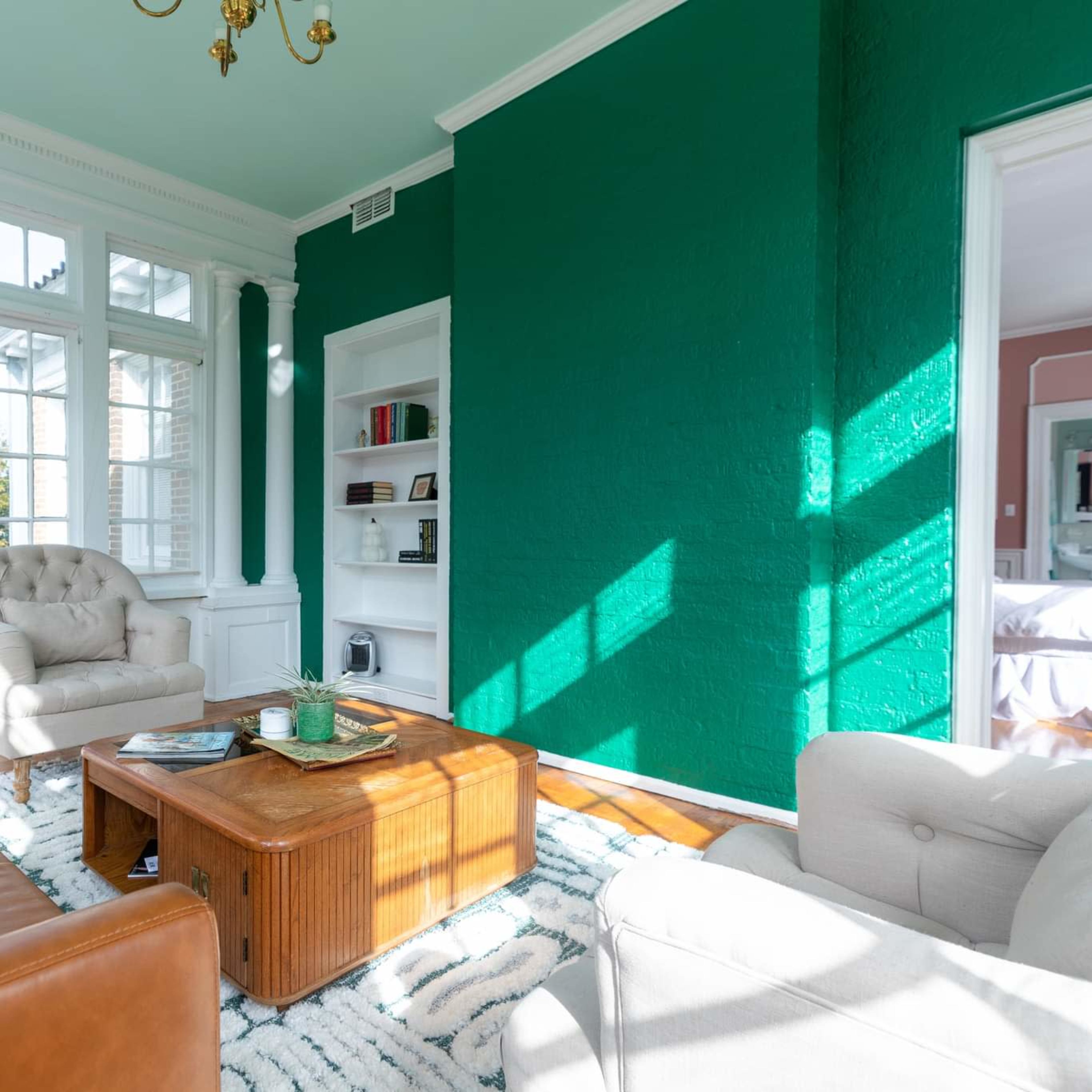 The room features a green accent wall, large windows allowing sunlight to stream in, and a mix of leather and fabric seating around a wooden coffee table.