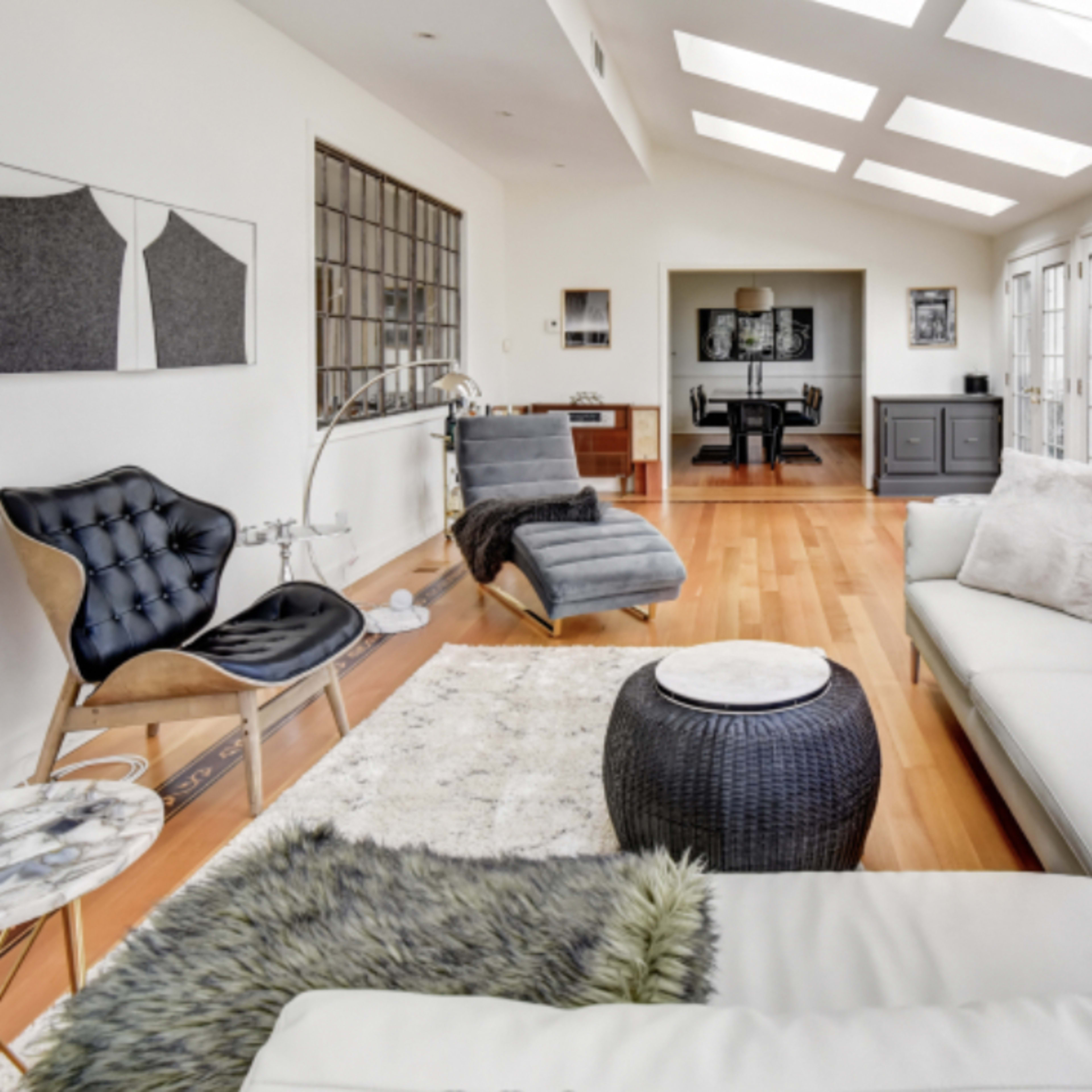A modern, bright living space featuring large windows, a white sectional sofa, and various pieces of stylish furniture arranged around a wooden floor.