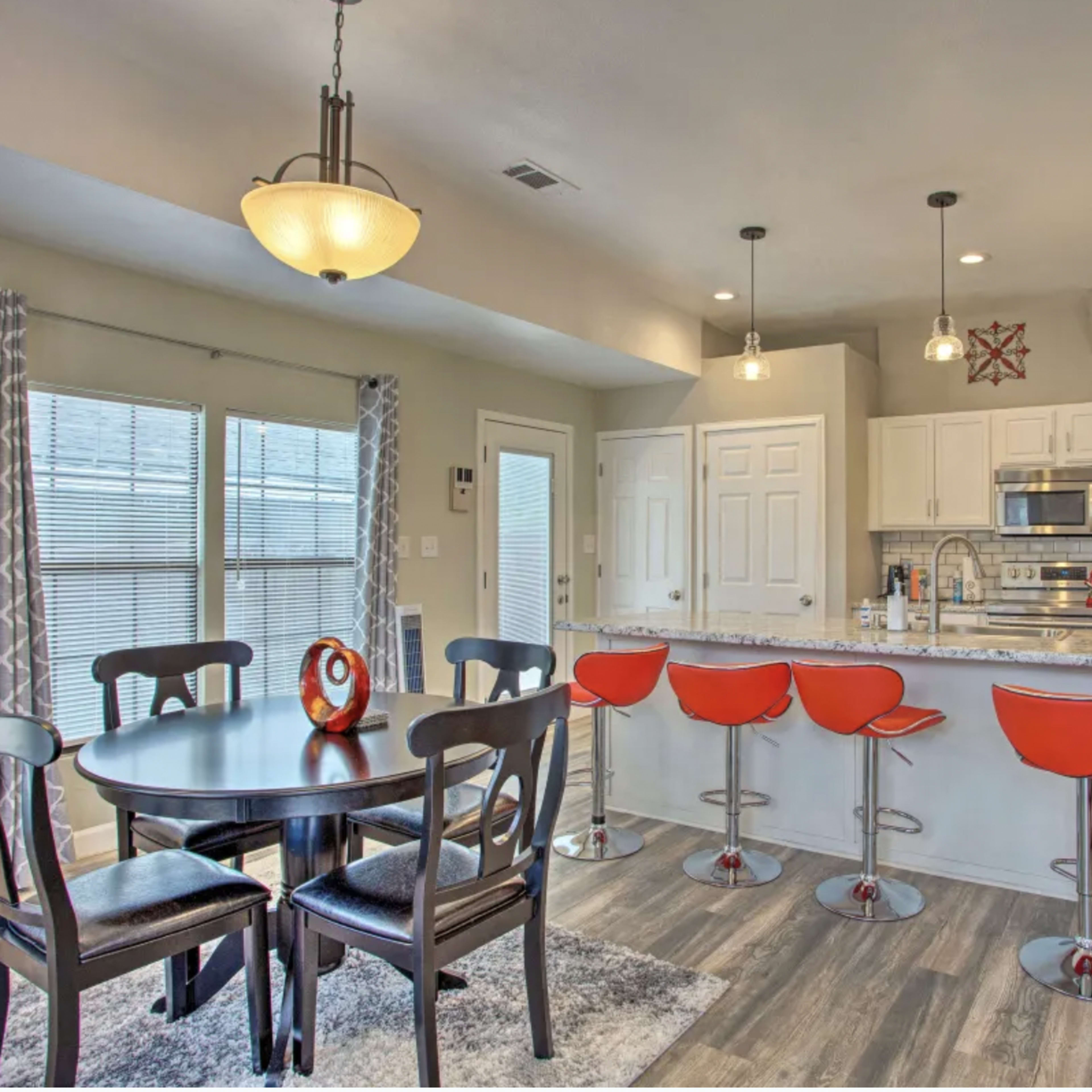 A modern kitchen with a dining area, featuring a round table with black chairs and red bar stools at a kitchen island, all set within a brightly lit space.