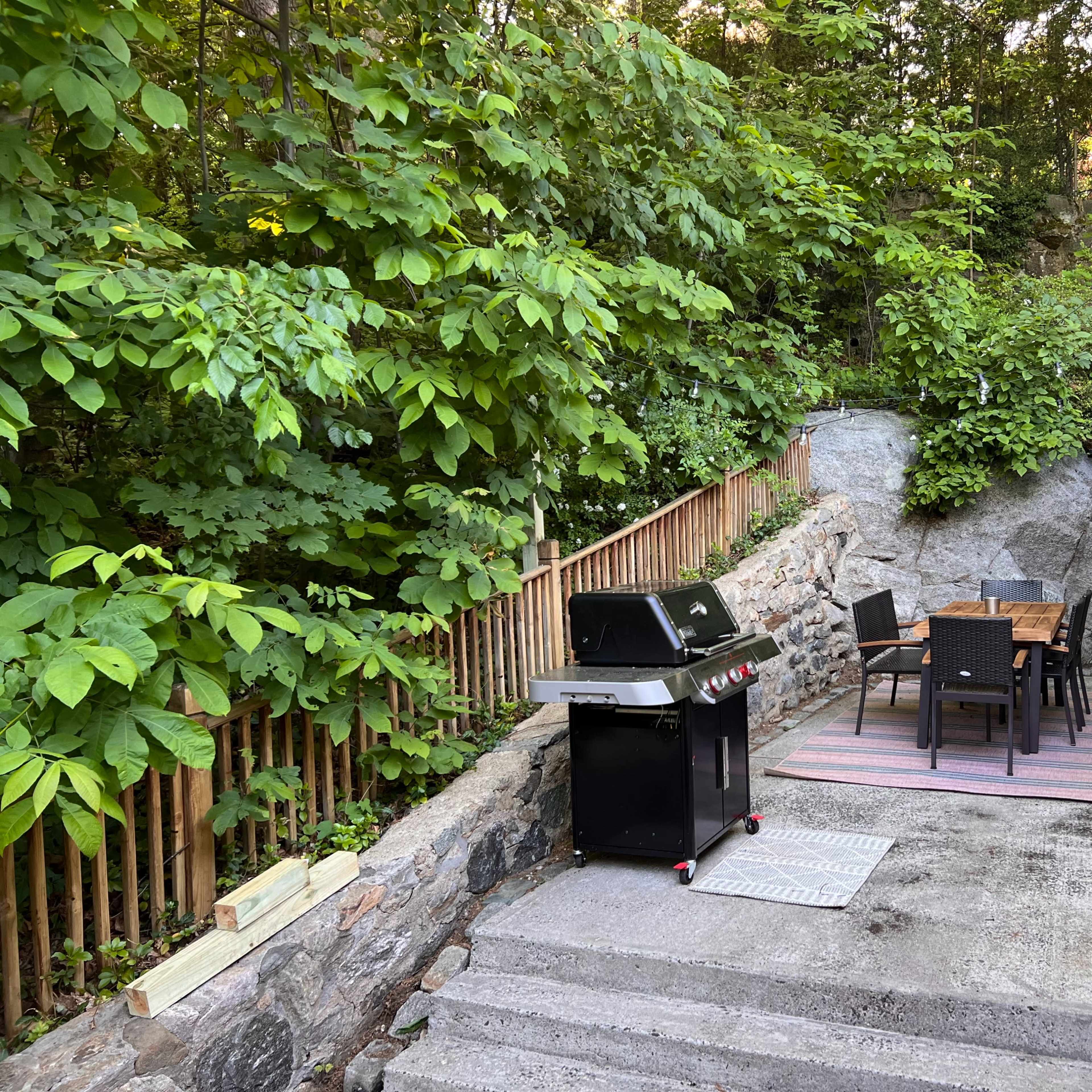 A patio area with a black barbecue grill and a dining table set with chairs, surrounded by lush greenery and stone walls.