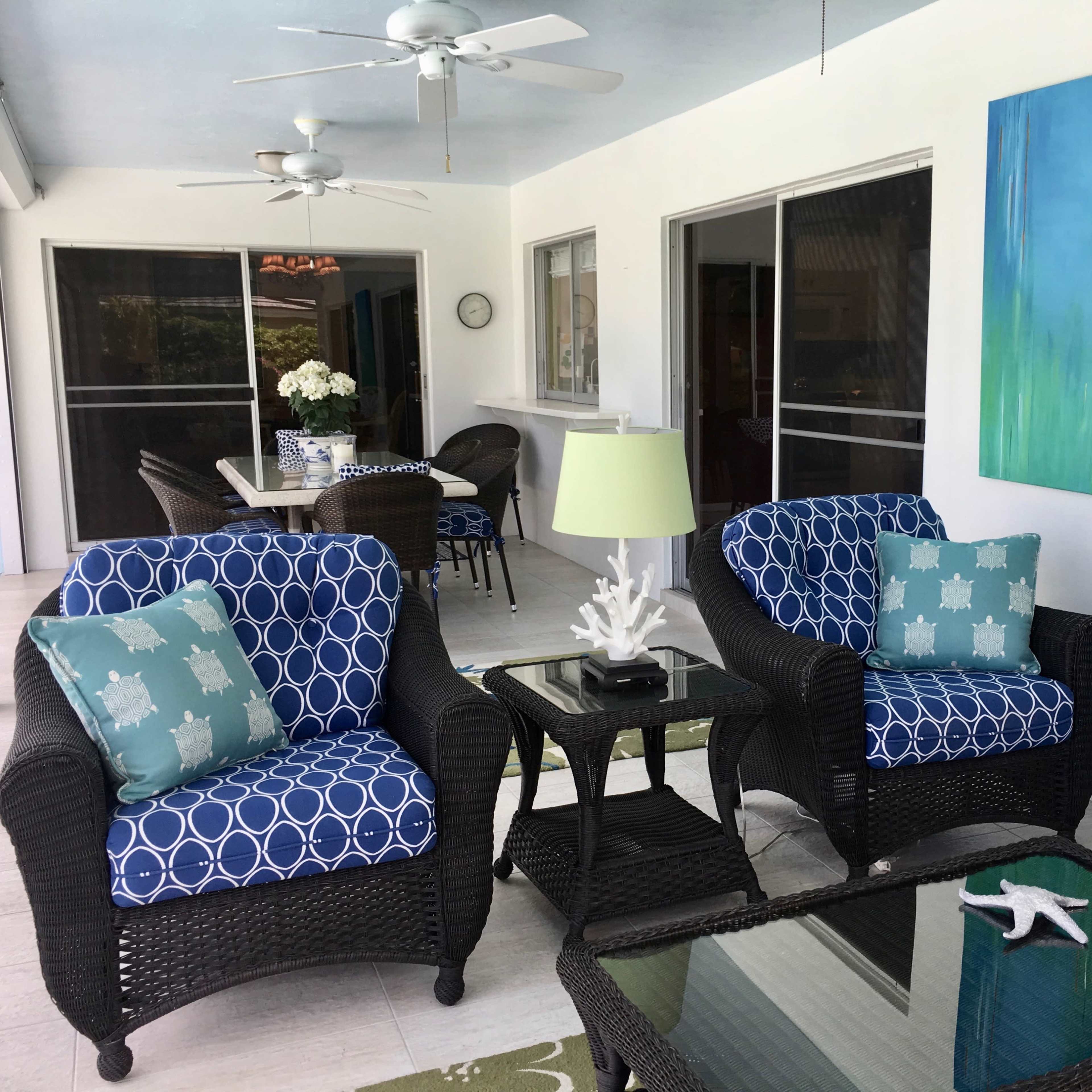 A porch features two patterned chairs with a small table between them, a dining area in the background, and a large blue painting on the wall.