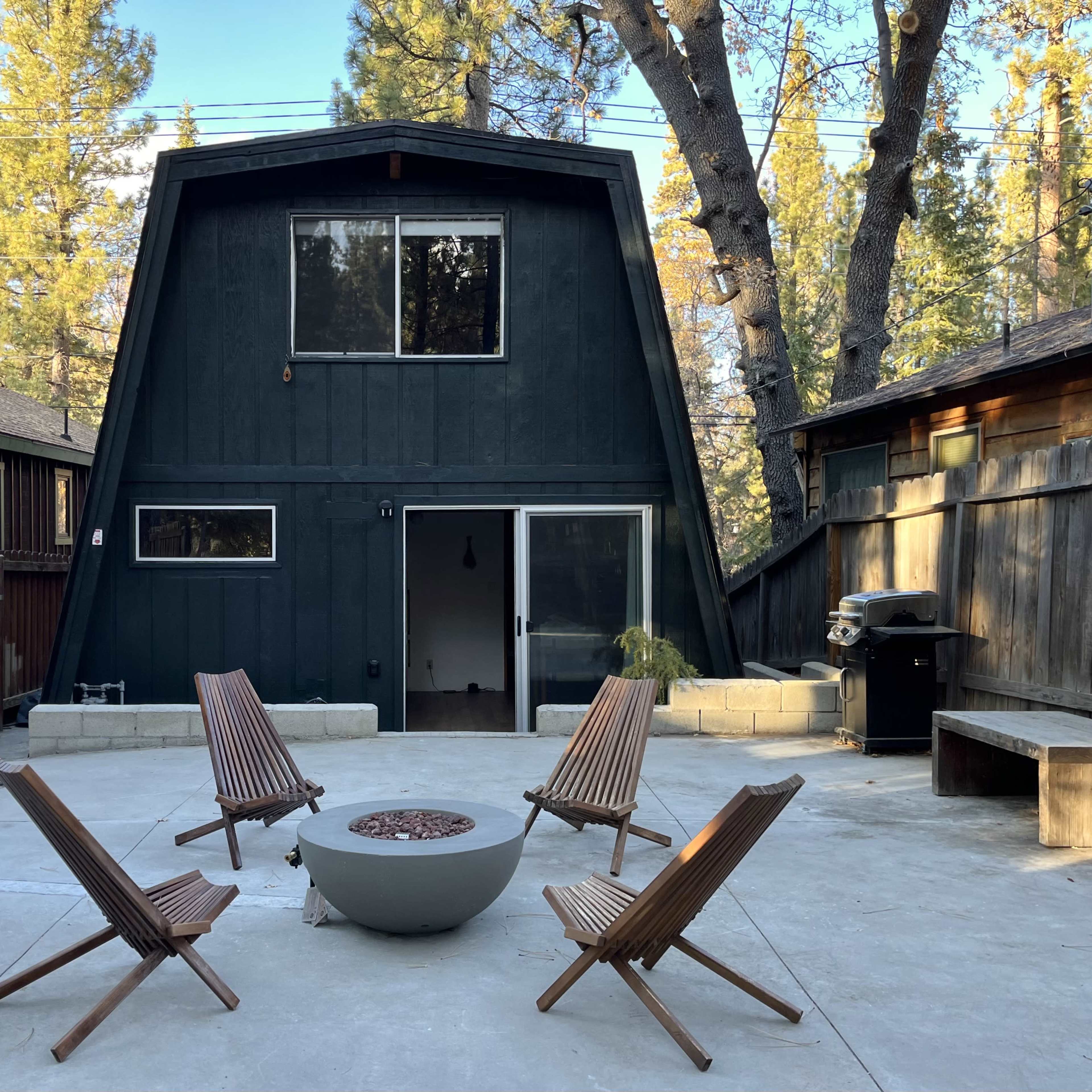A modern A-frame house sits in a forested area, surrounded by gravel and wooden chairs arranged around a fire pit.