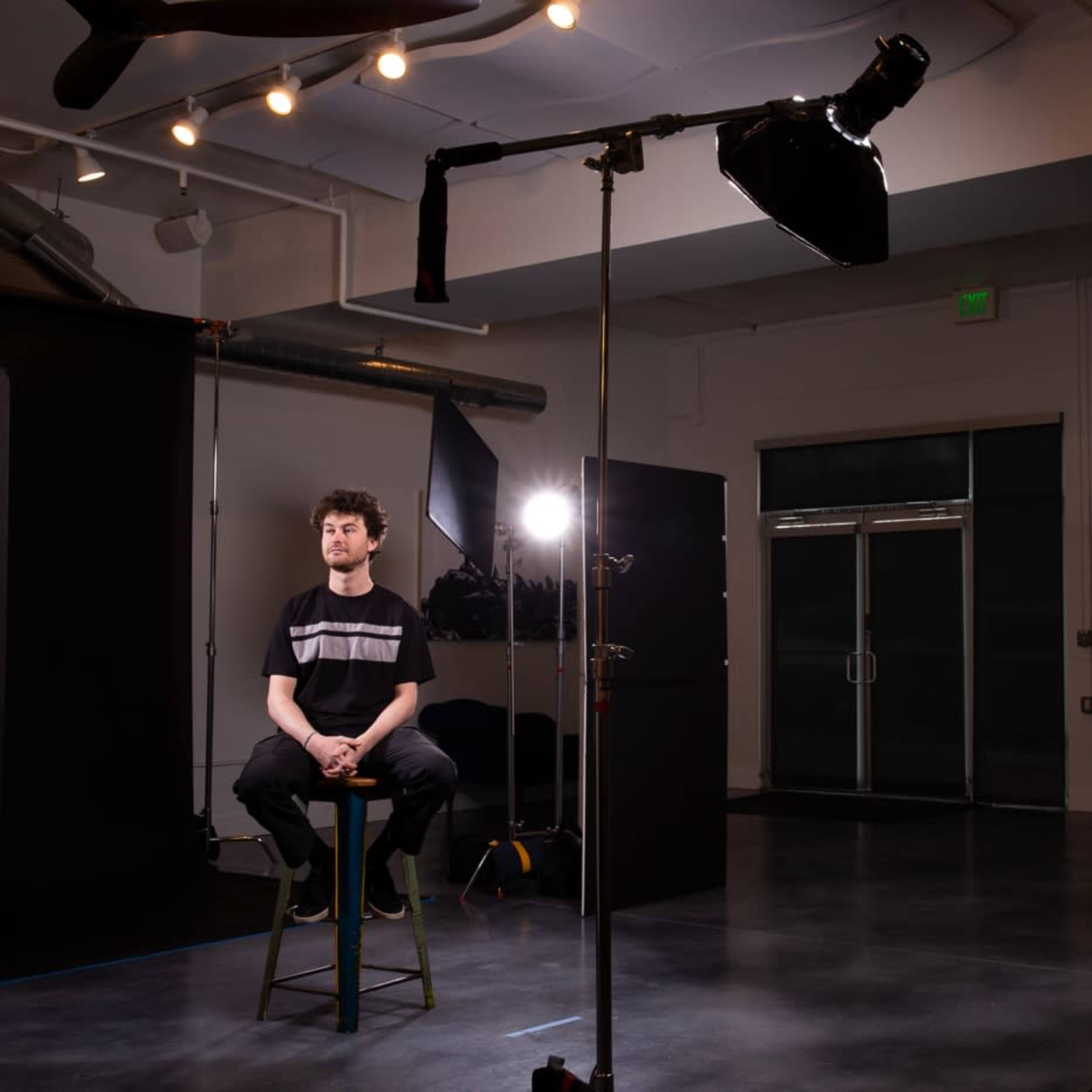 A young man sits on a stool in a photography studio, surrounded by backdrops and studio lights.