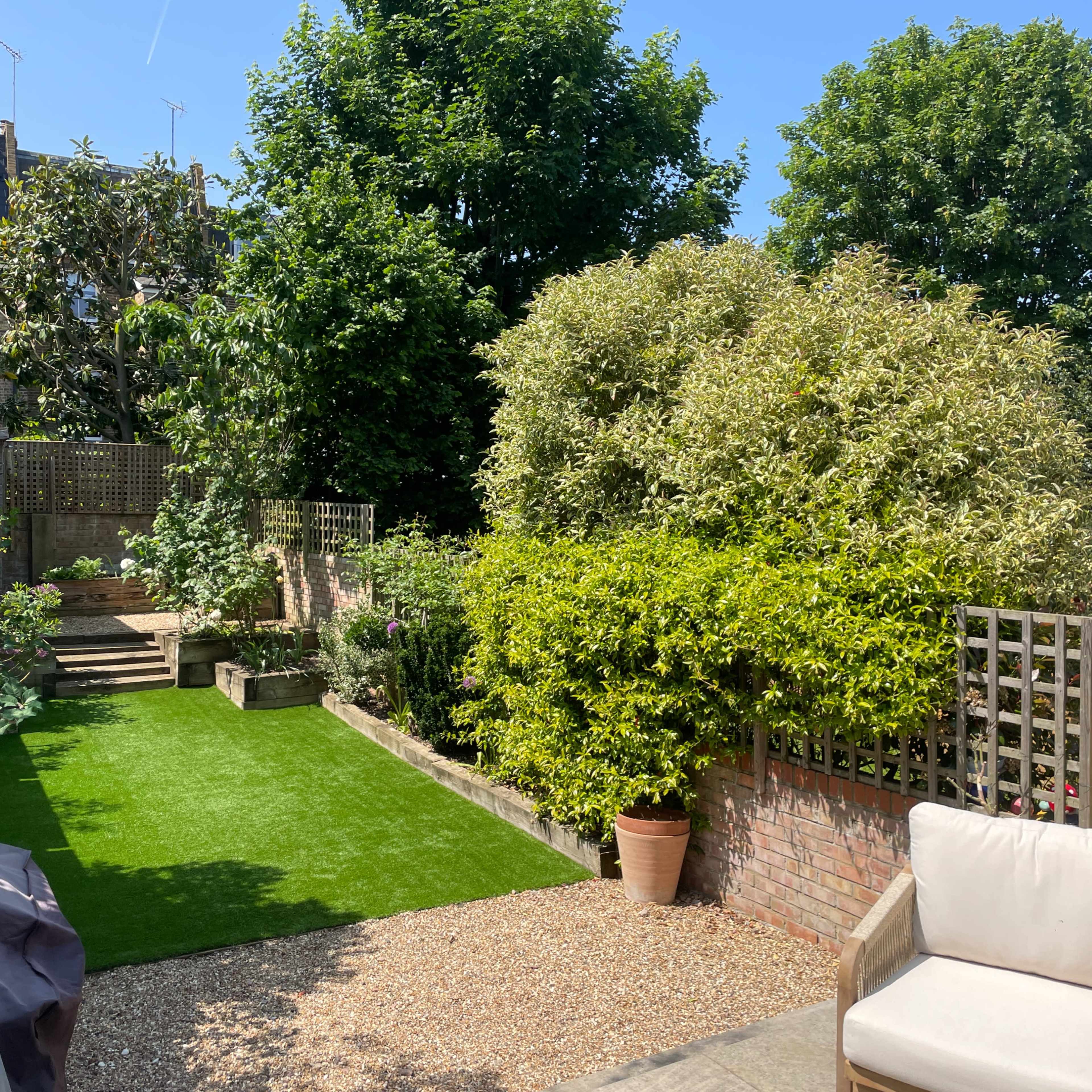 A landscaped garden with green grass, stone steps, a gravel area, potted plants, and a chair under a clear blue sky.