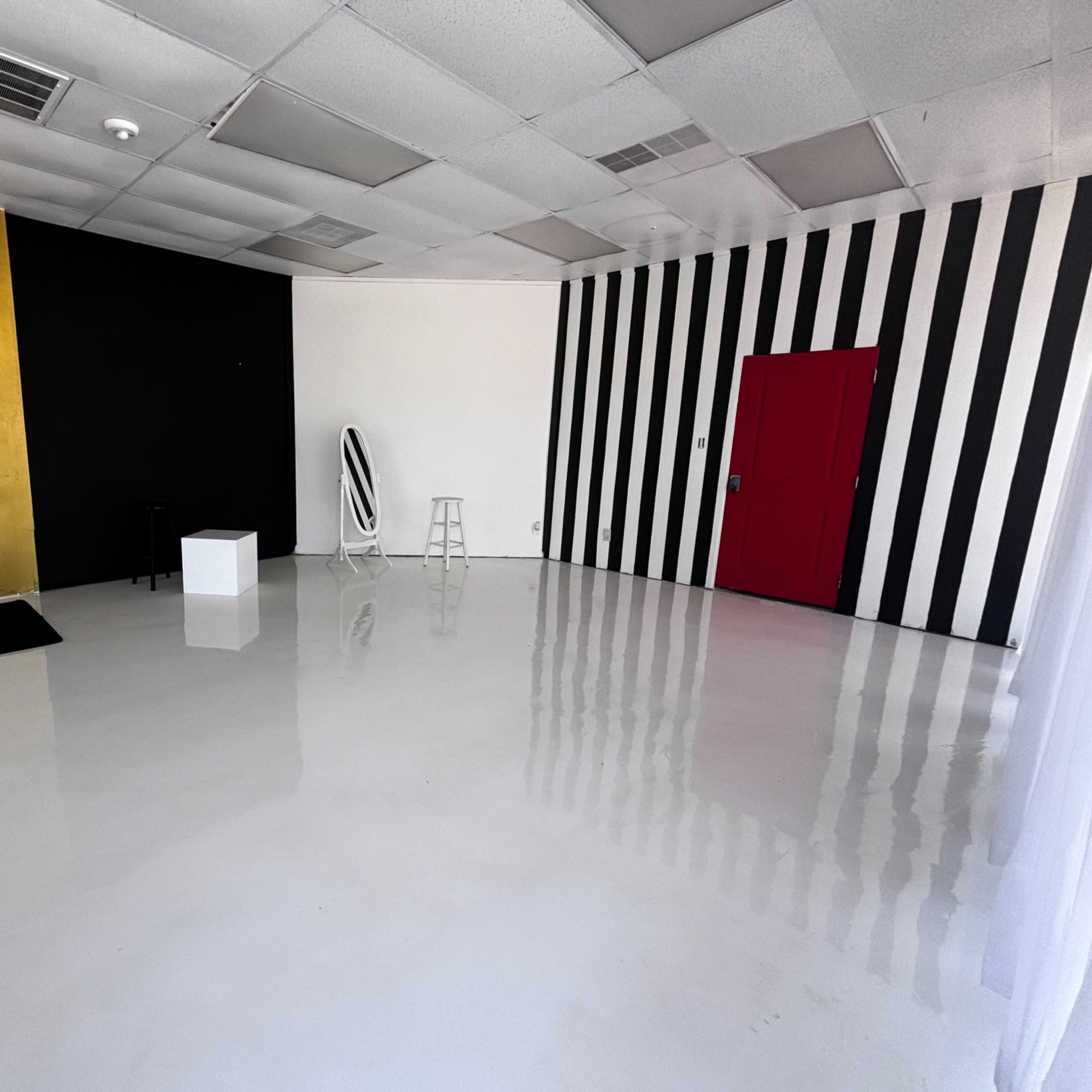 An empty room with a shiny white floor, a gold and black wall, a red door, and black and white striped walls.