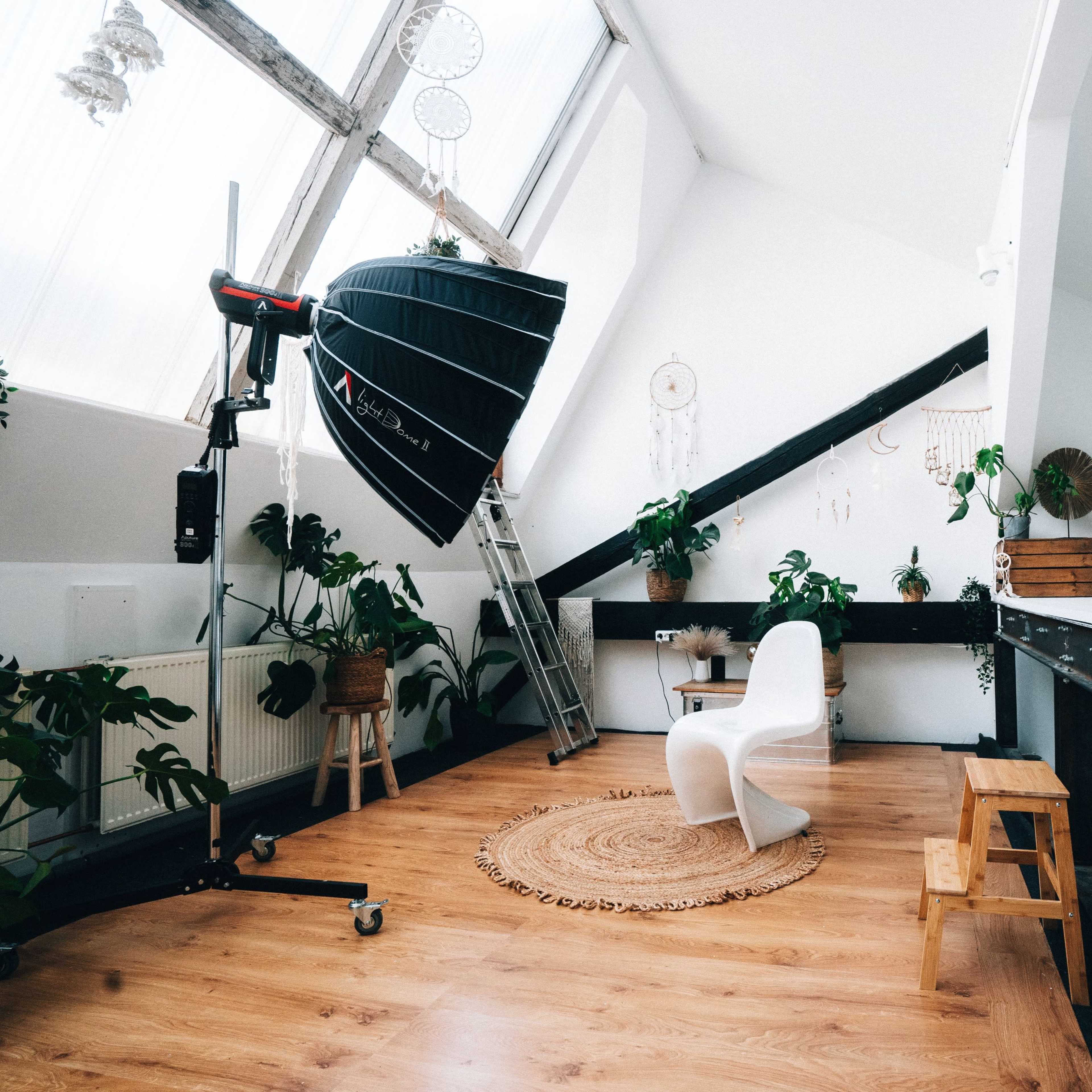 A well-lit attic studio with wooden flooring, a white chair, a large photography light, and various plants along the walls.