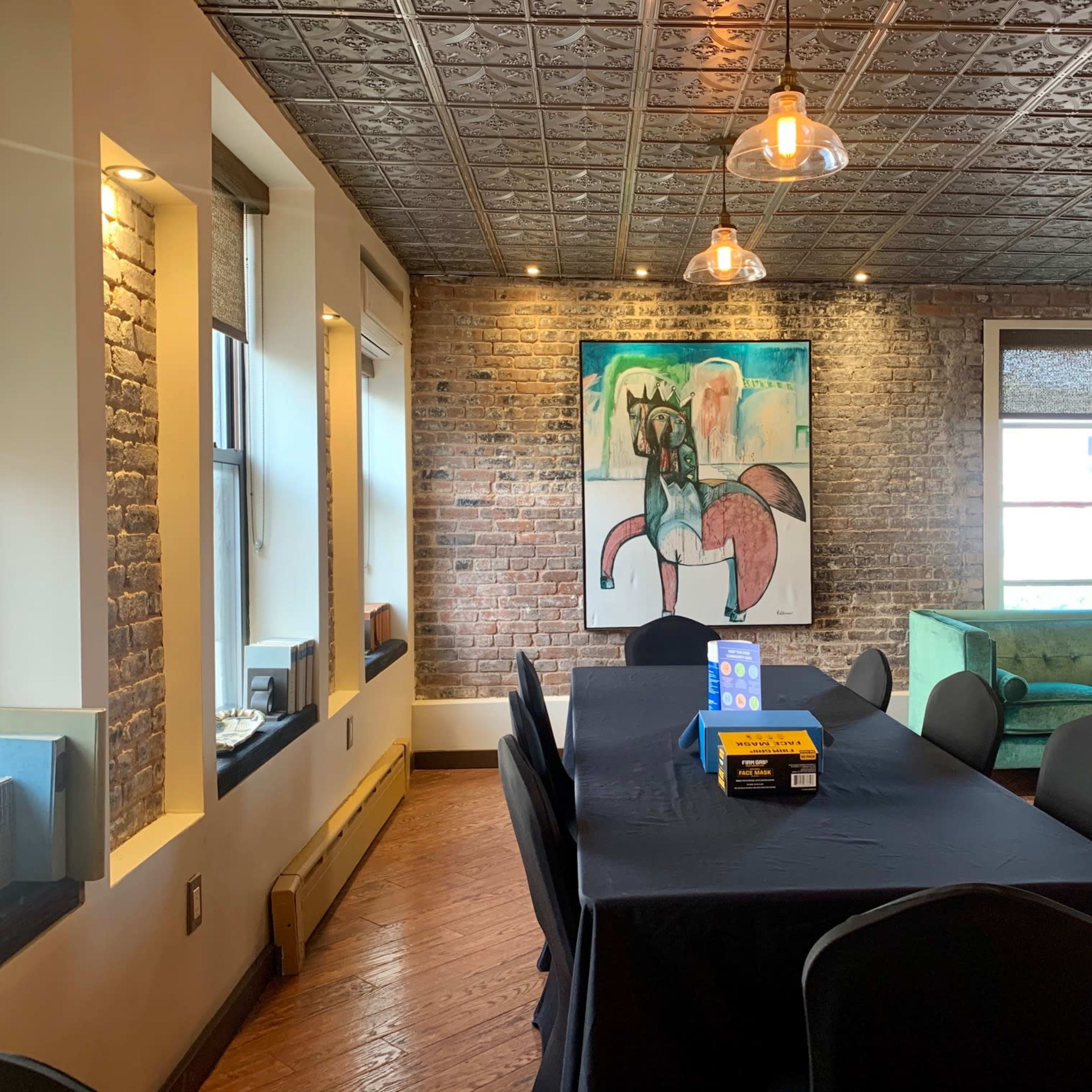 A modern meeting room with a large black table, black chairs, a green sofa, exposed brick walls, and a colorful artwork on the wall.