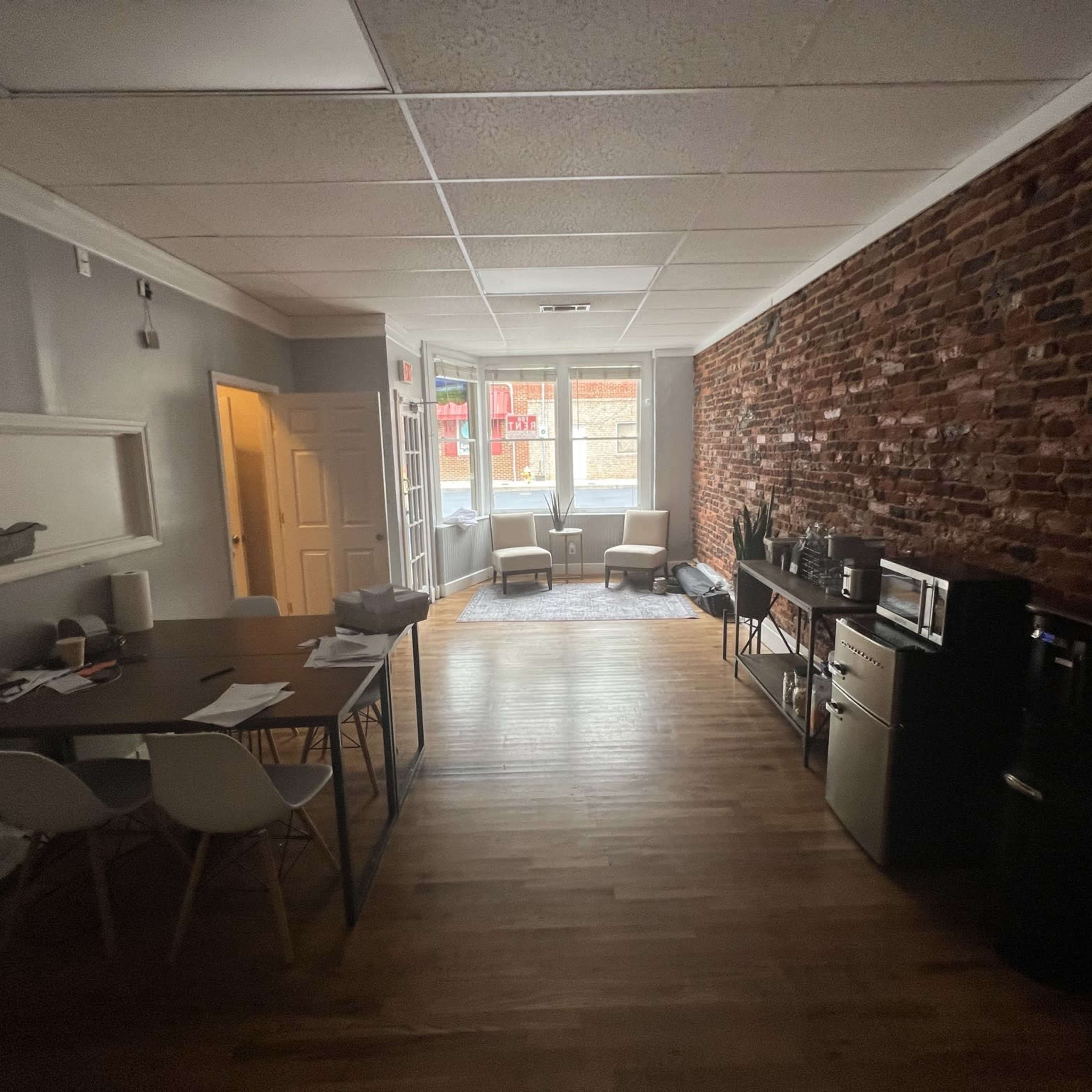 A spacious room with exposed brick walls, hardwood flooring, a dining table with chairs, and two armchairs near a window.