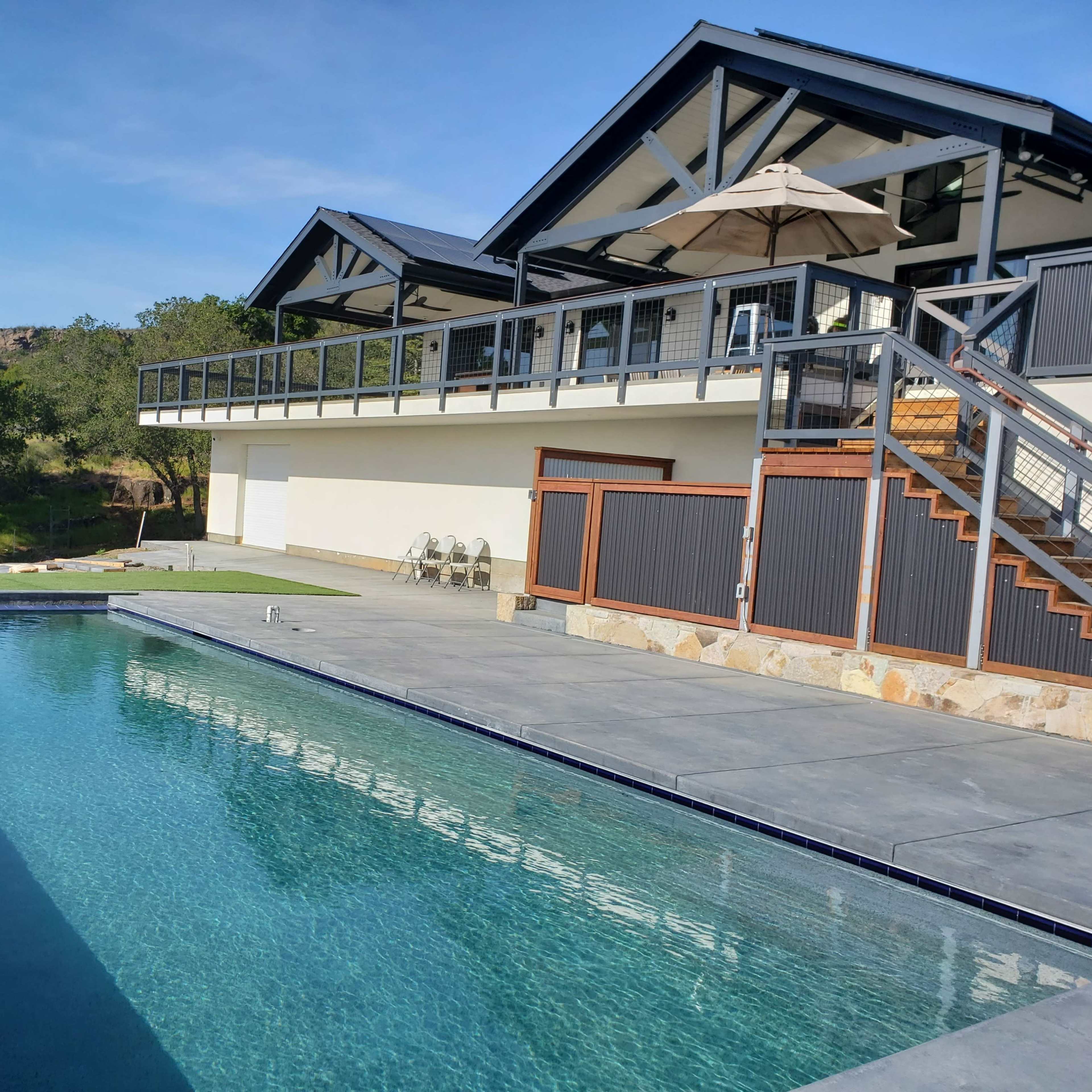 A modern two-story house with a deck overlooks a rectangular swimming pool surrounded by stone and concrete.