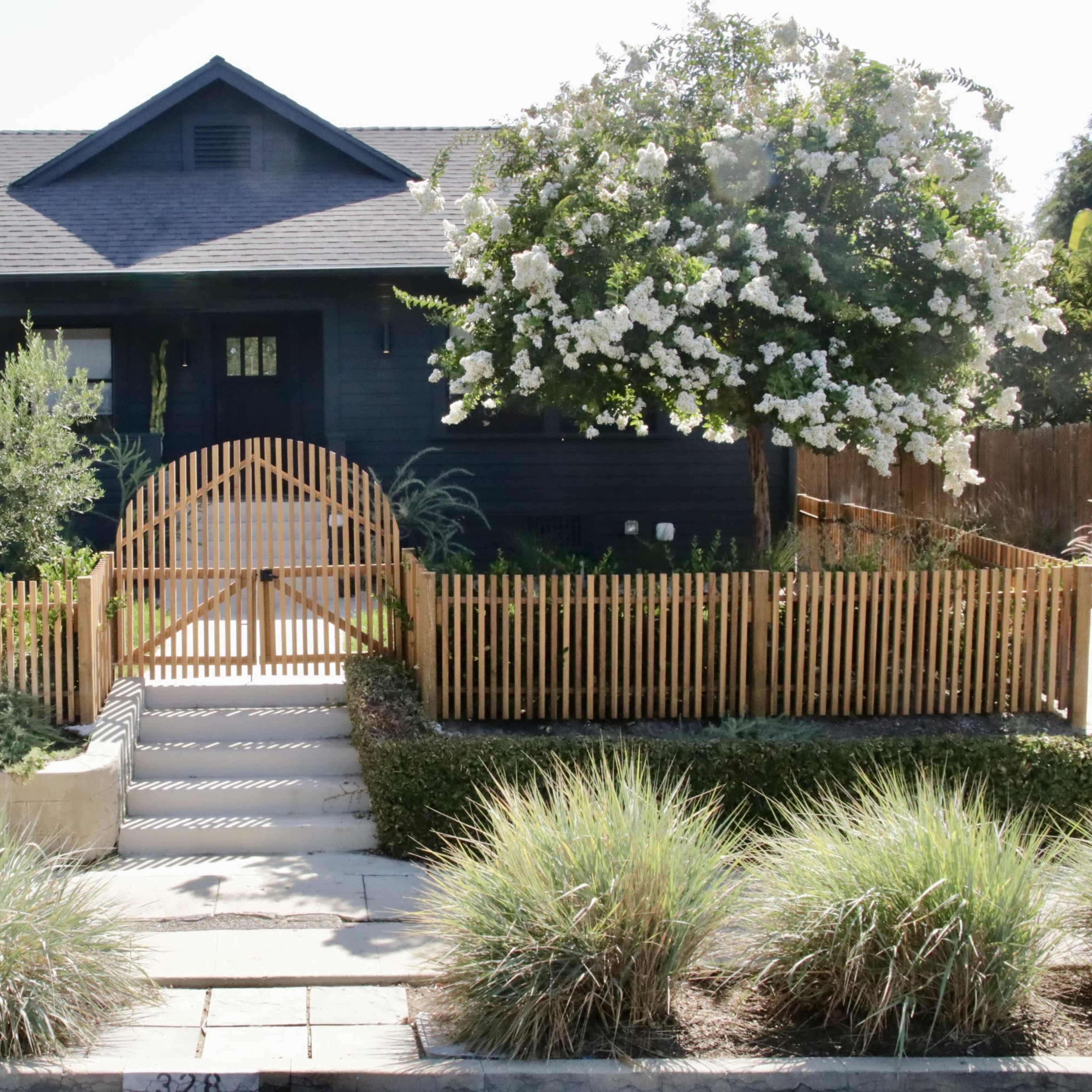 A blue house with a front porch is surrounded by a wooden fence and a garden, featuring a flowering tree.