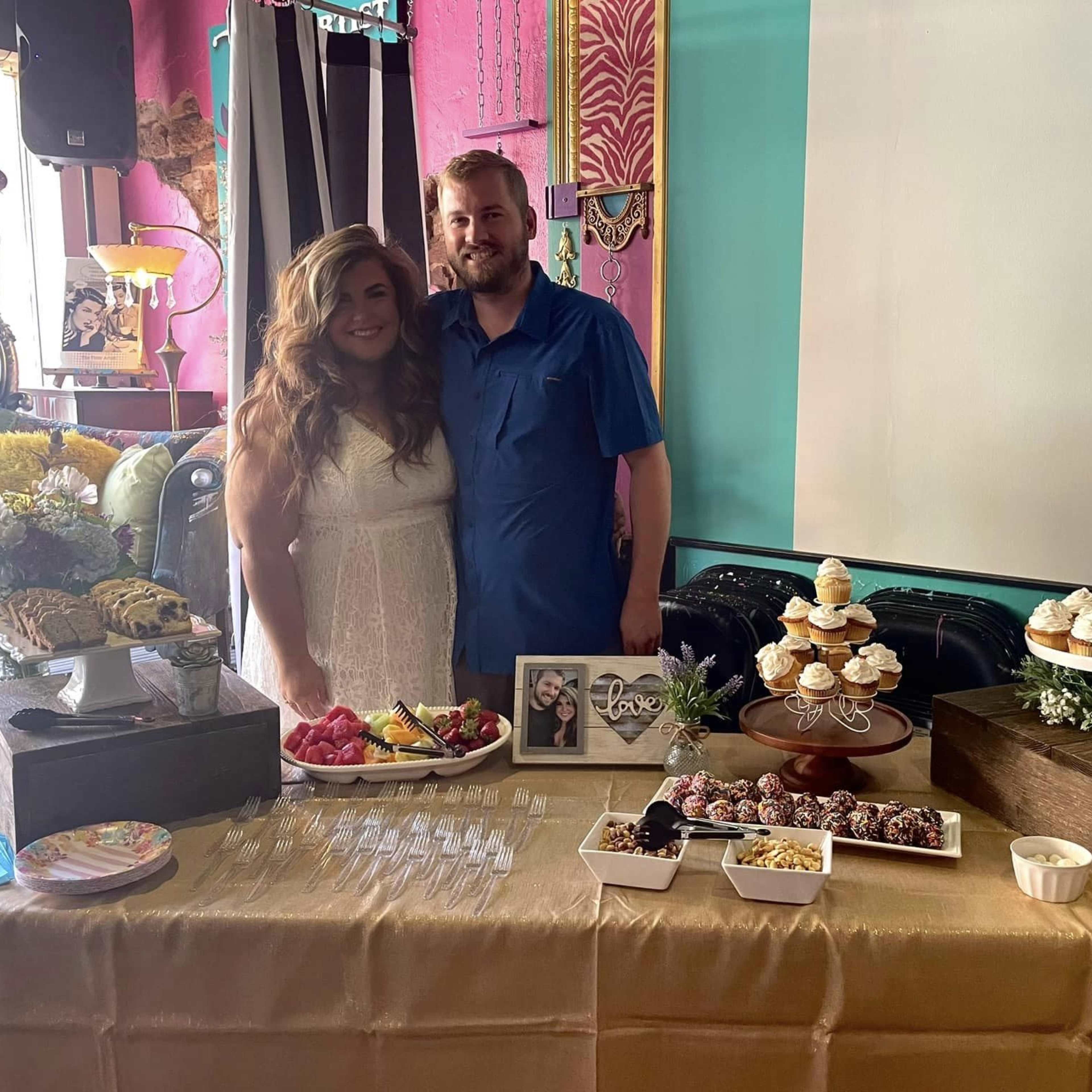 A couple stands together behind a table adorned with various desserts and floral arrangements, set in a colorful indoor space.