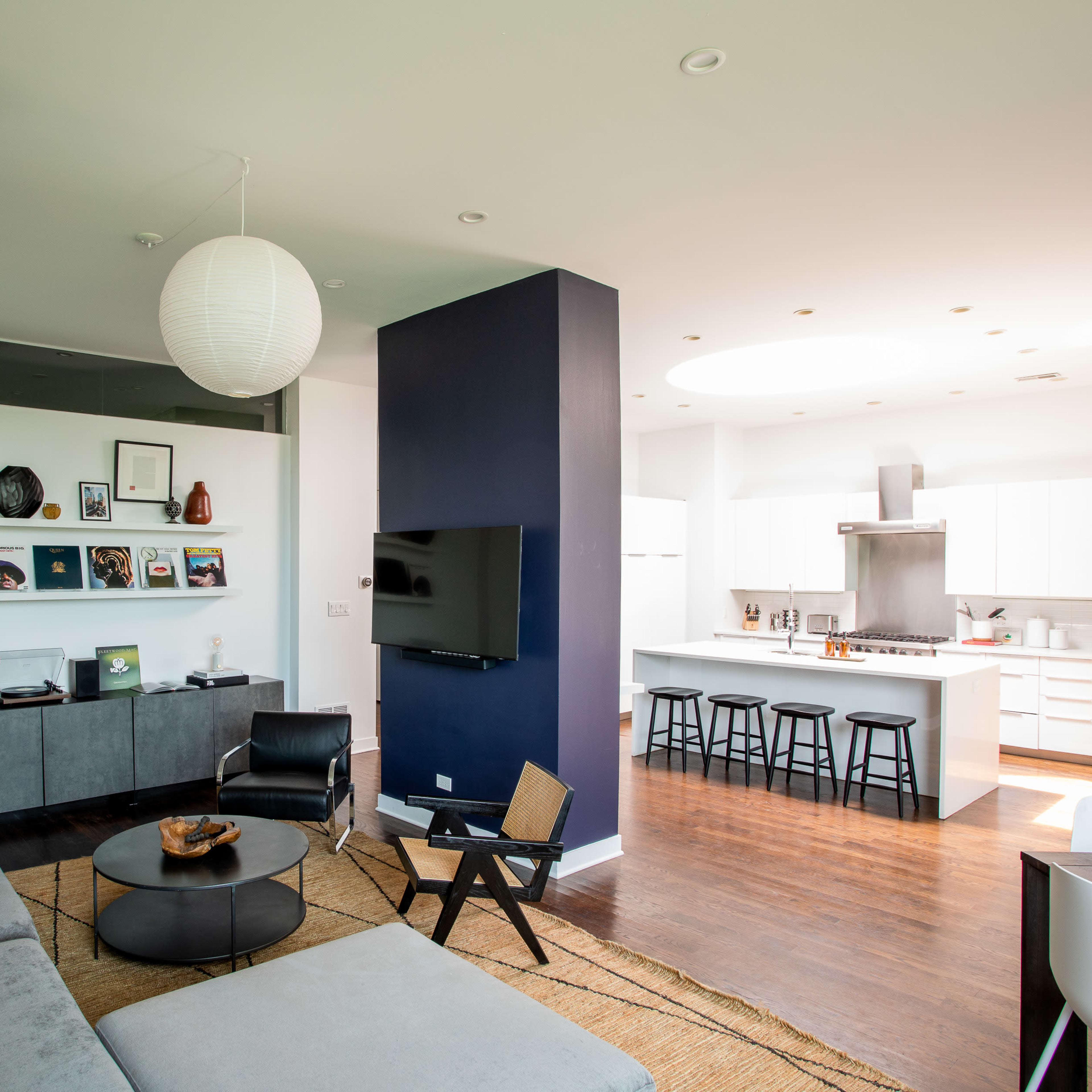 A modern living space features a gray sofa, a round coffee table, a television mounted on a blue wall, and an adjacent kitchen with white cabinetry and bar stools.
