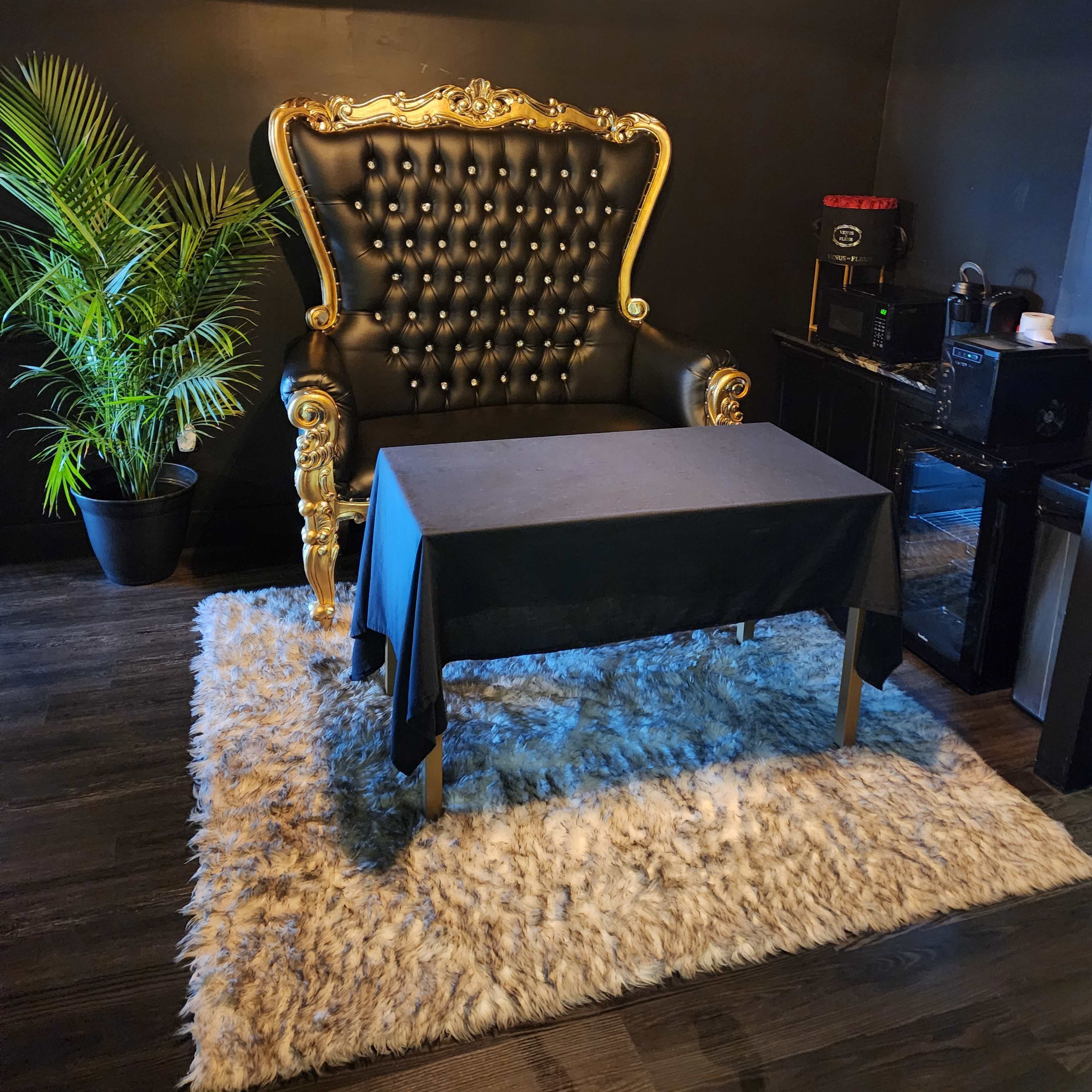 A black and gold throne-style chair next to a small table on a fuzzy rug, with a plant in a pot nearby and a kitchenette in the background.