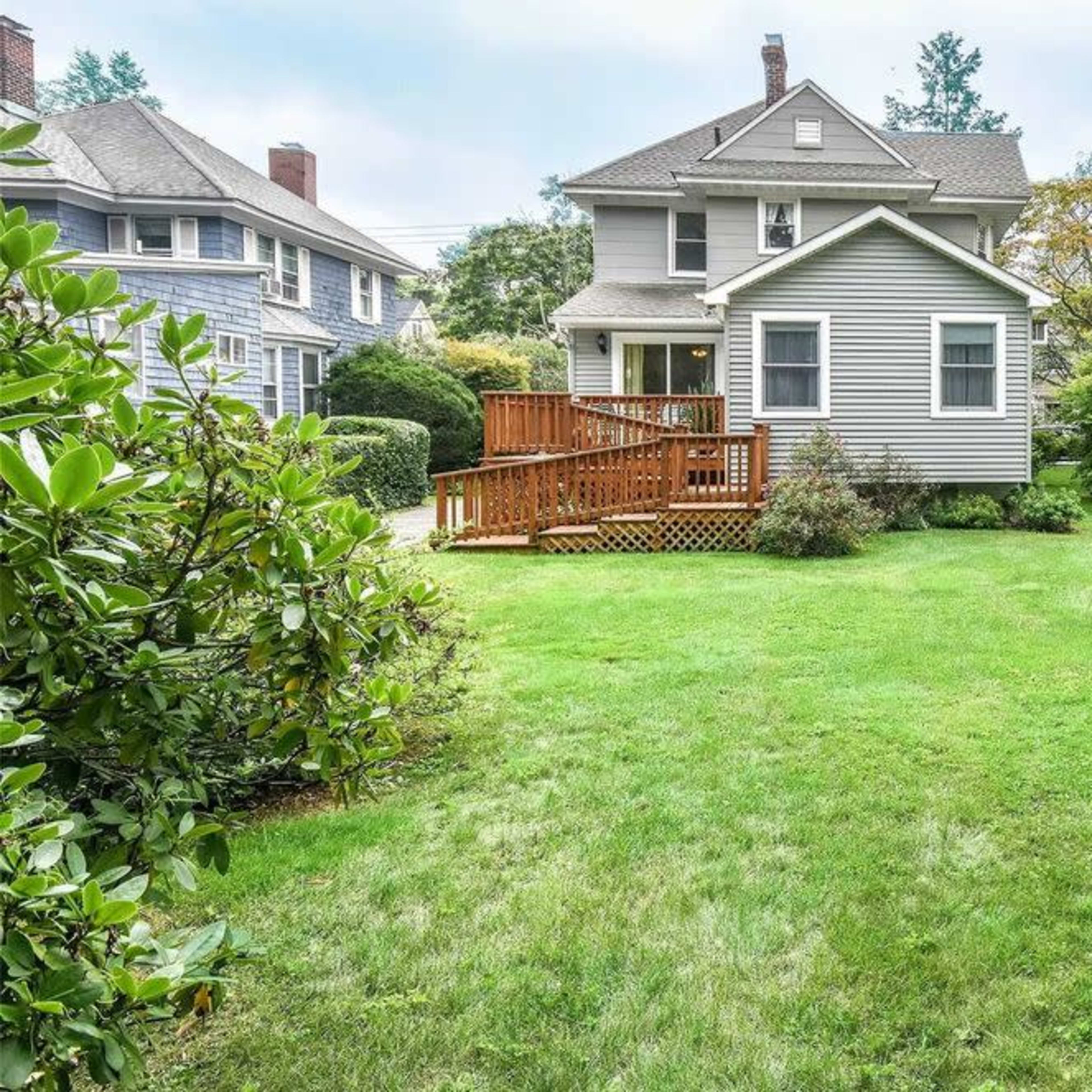 A residential yard with a two-story house, a wooden deck, and well-maintained grass surrounded by shrubs and trees.