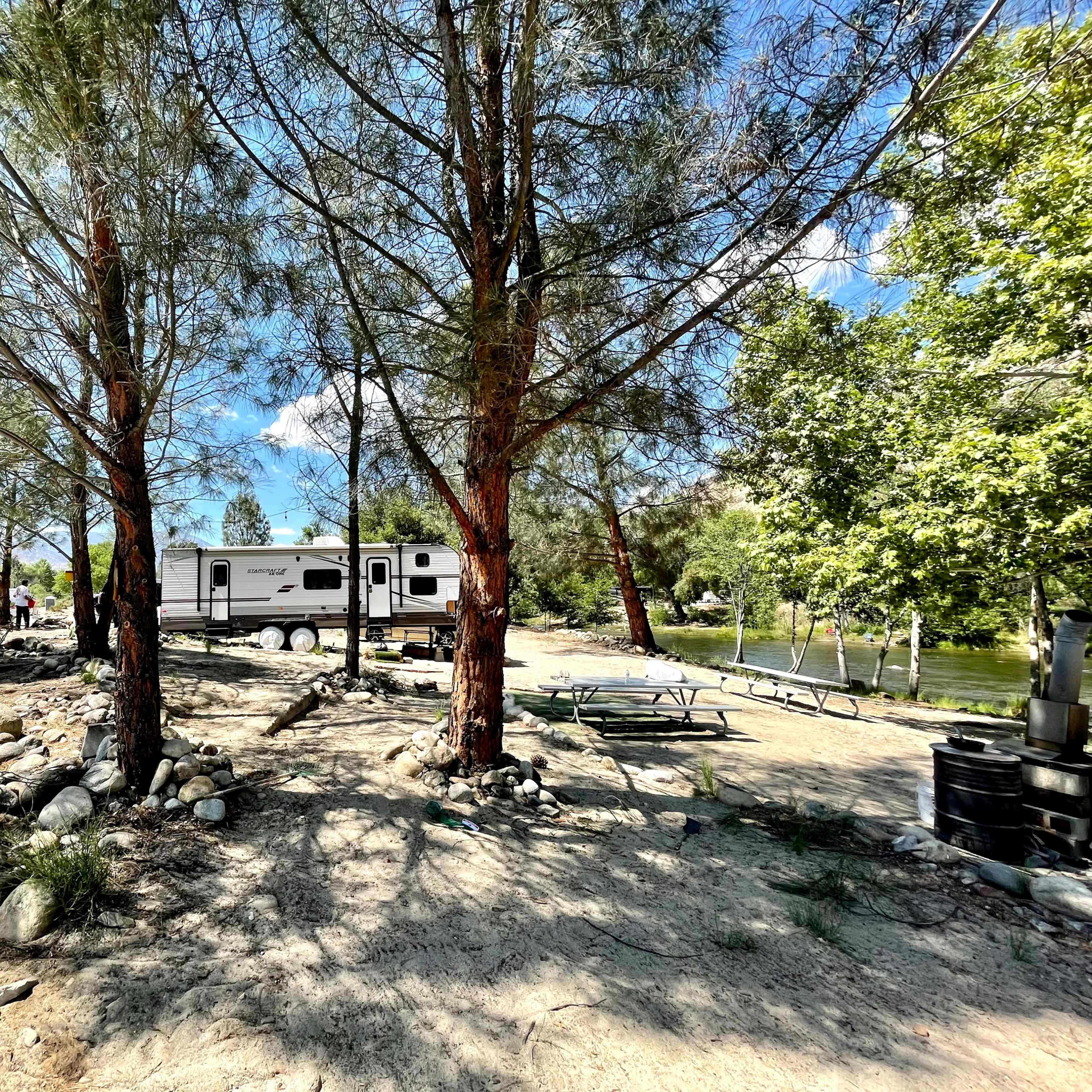 A recreational vehicle is parked among trees near a river, with a picnic area and boulders in the foreground.