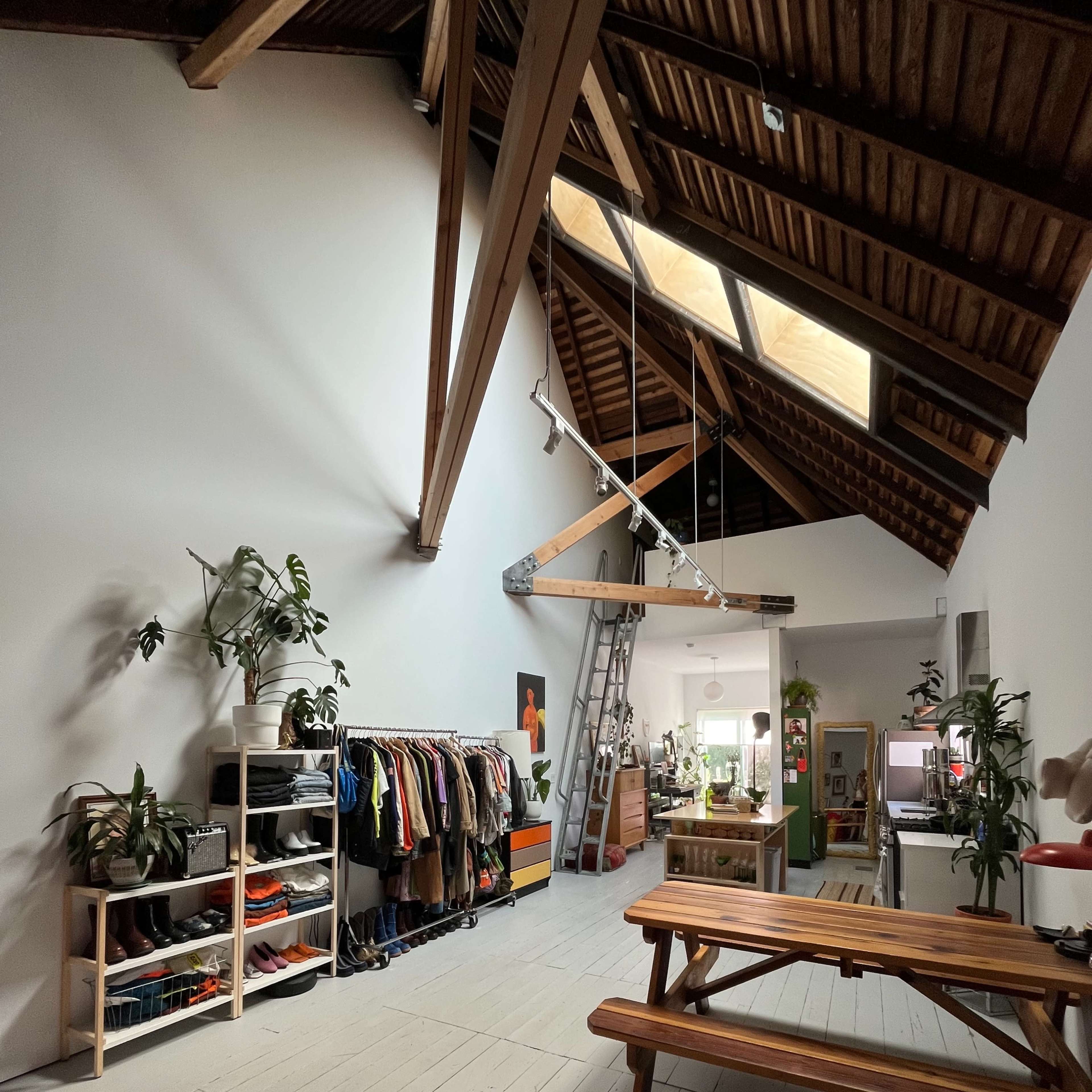 A spacious interior with wooden beams, a ladder, and organized shelving displaying clothing and plants alongside a picnic table.