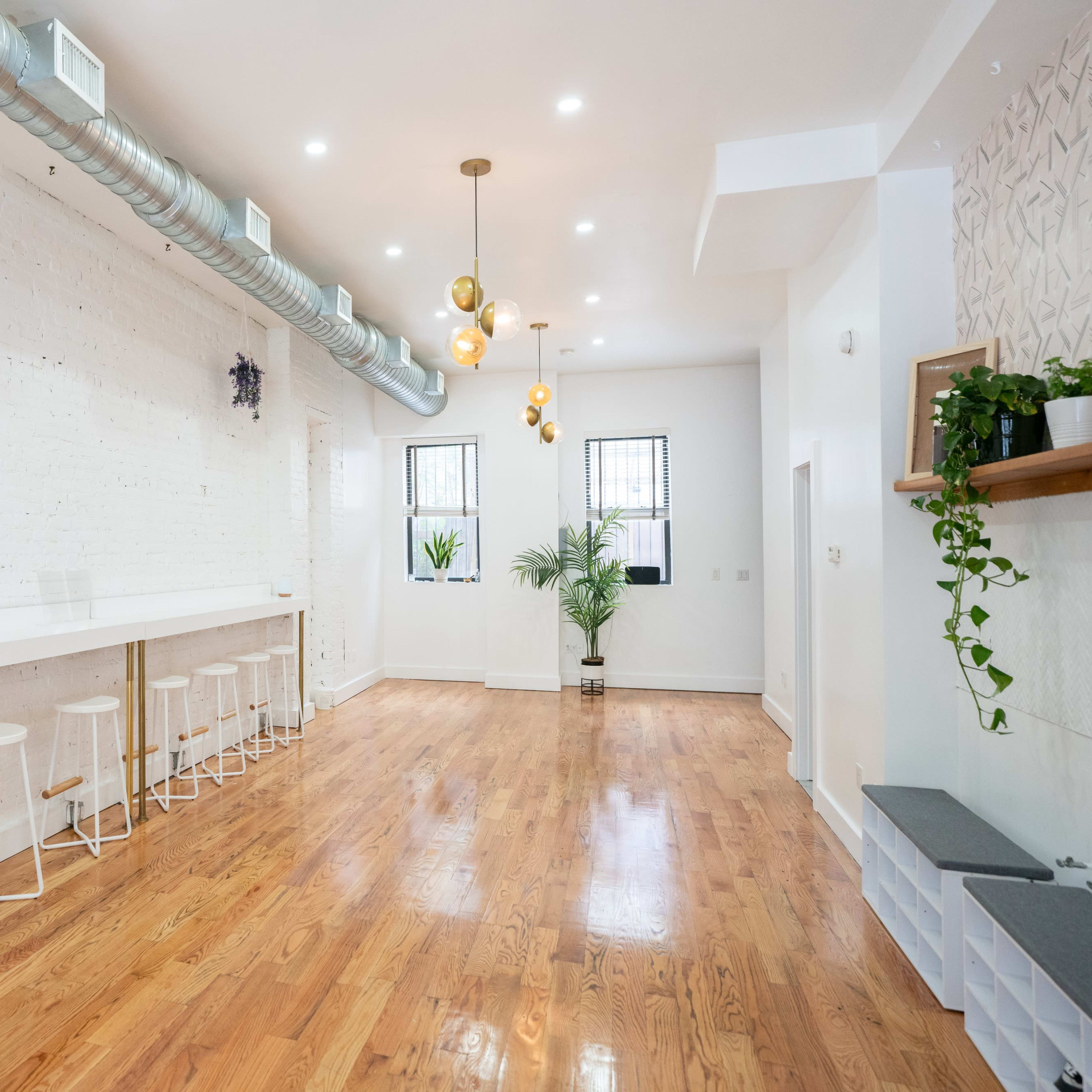 A spacious interior with white walls, wooden floors, a long bar area with white stools, and several plants.