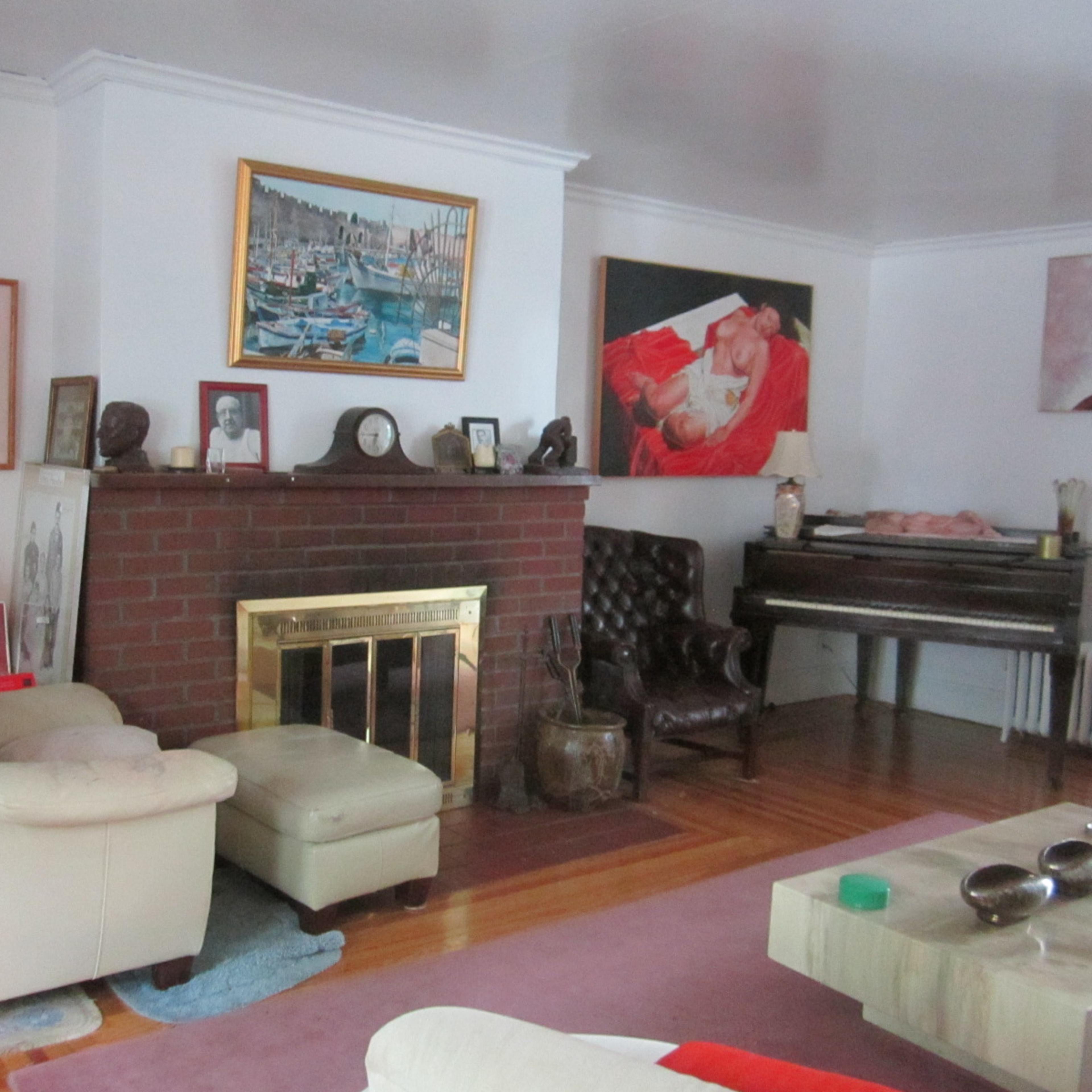 A cozy living room with a brick fireplace, a piano, and various artworks on the walls.