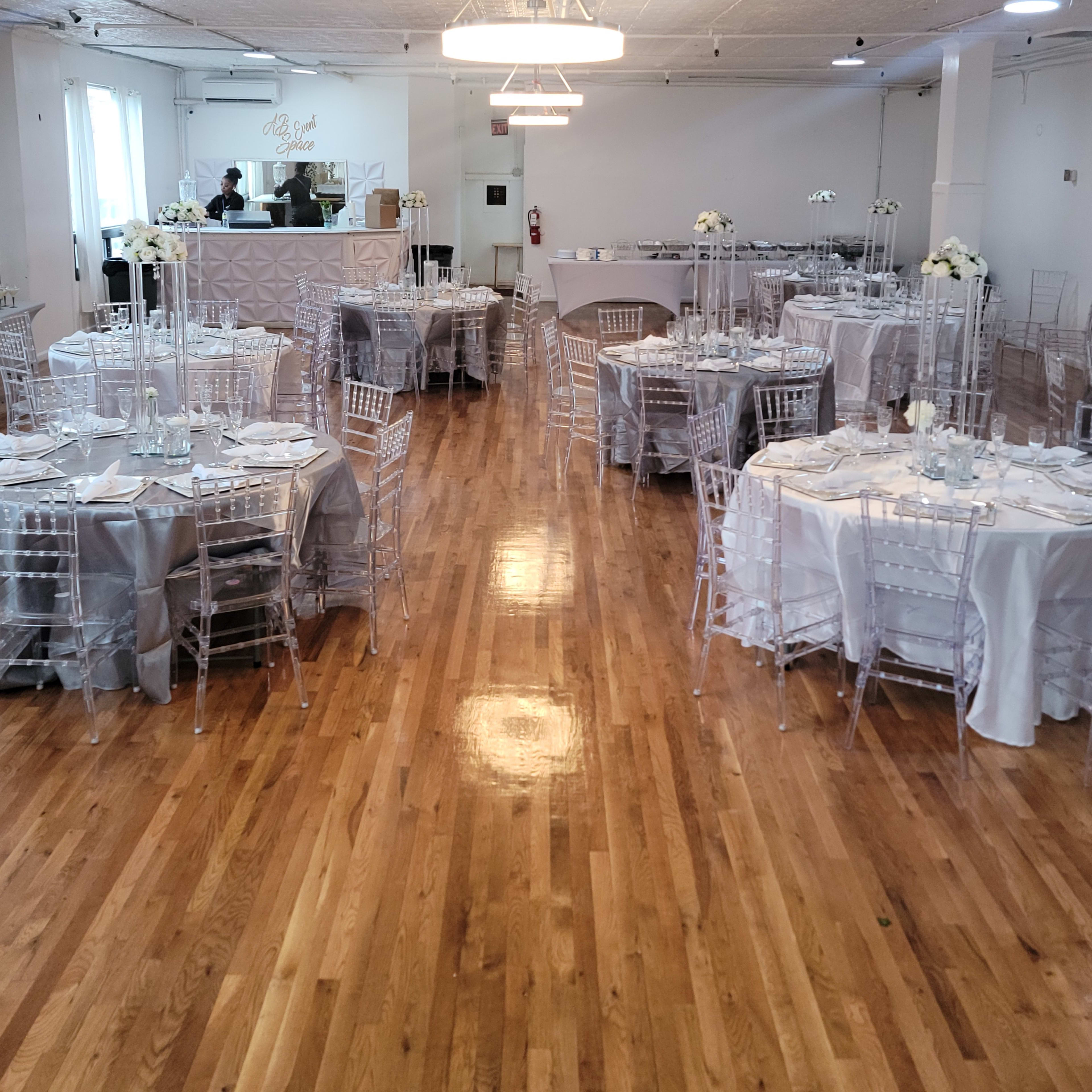 A spacious event room is set up with tables covered in silver linens and adorned with centerpieces, featuring clear chairs and polished wooden floors.