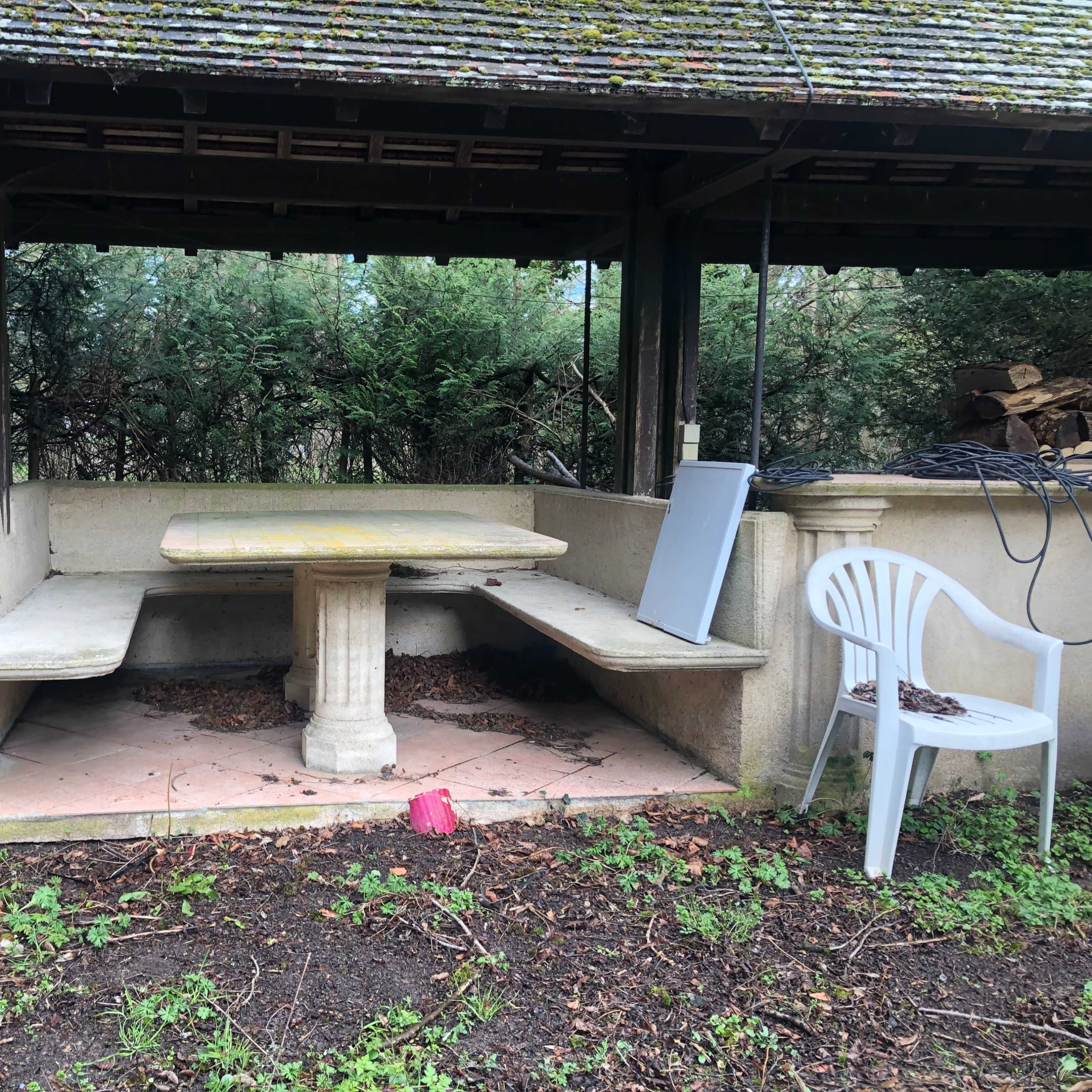 A partially covered outdoor seating area with a stone table and benches, surrounded by greenery.