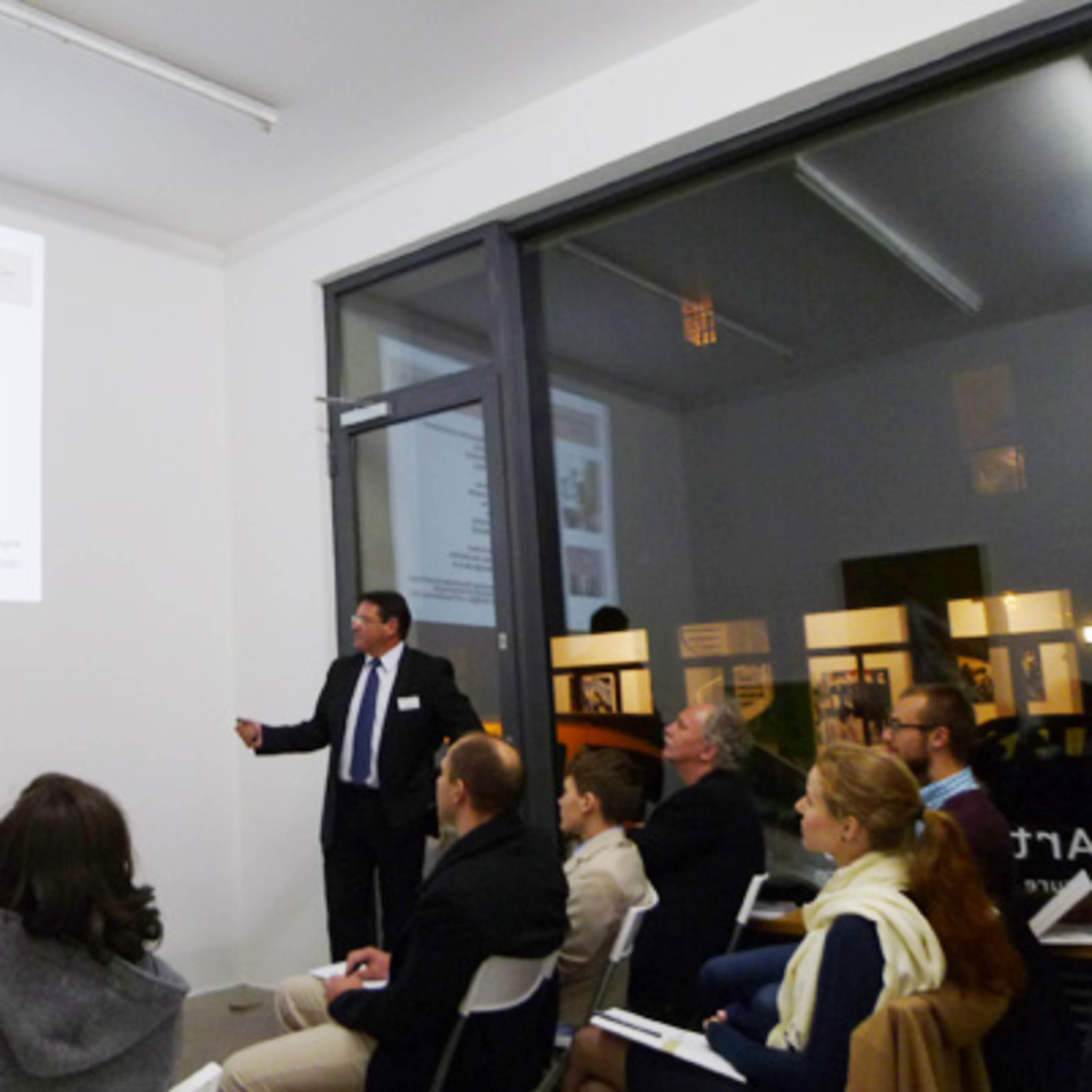 A presenter in a suit points toward a slideshow while an audience observes in a well-lit room.