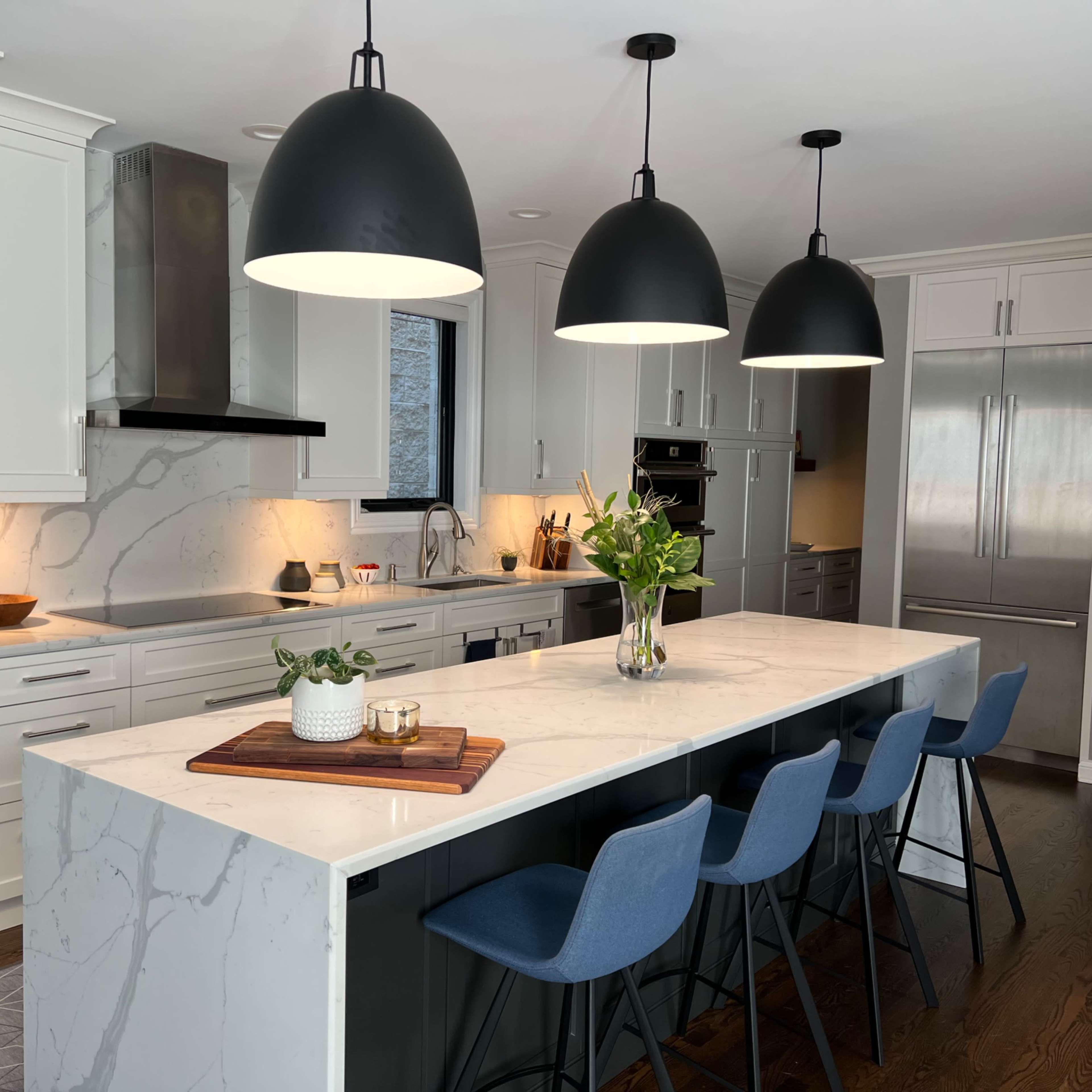 A modern kitchen features a large island with a marble countertop and blue bar stools, illuminated by three black pendant lights.