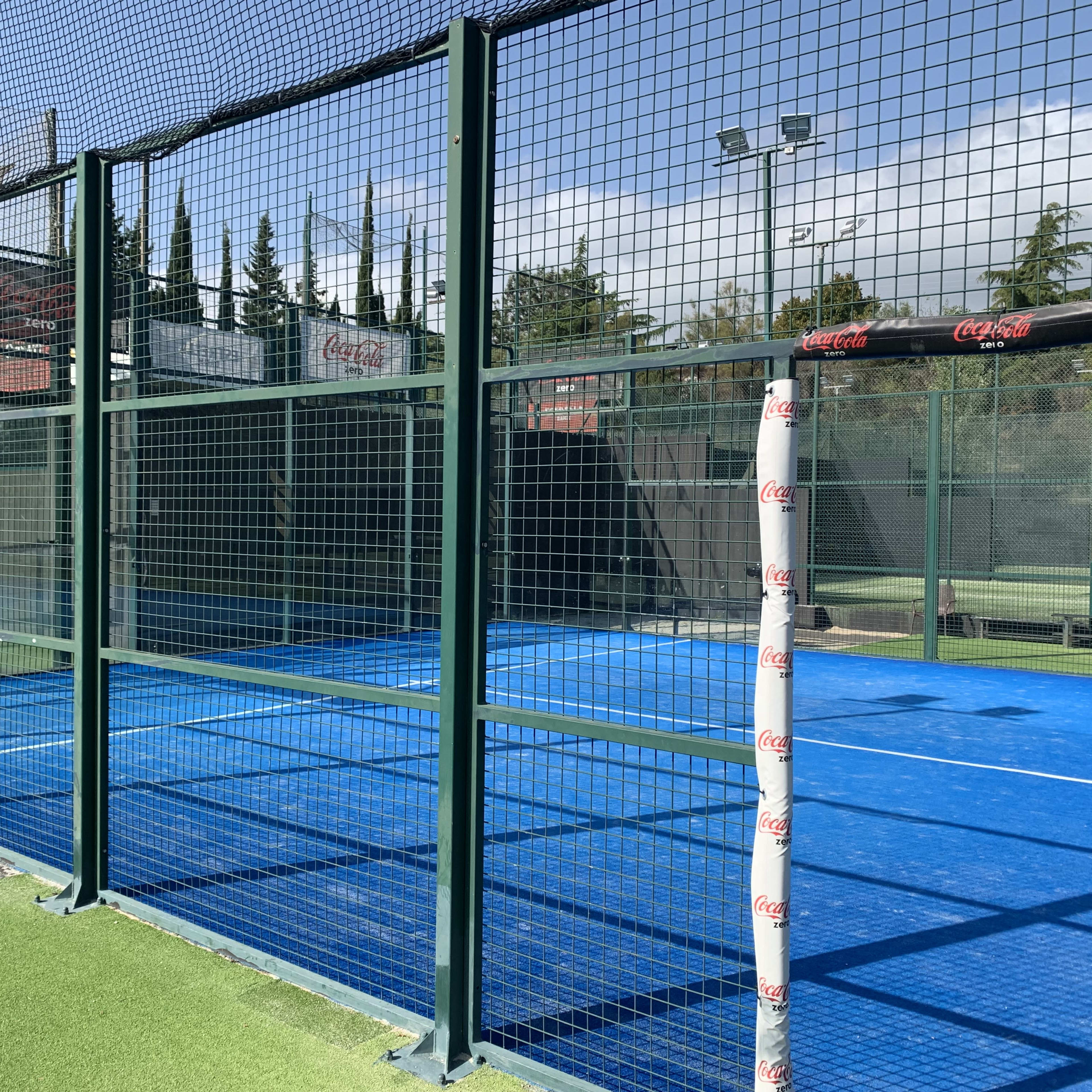 A blue padel court enclosed by green metal fencing, with a visible net and surrounding facilities.
