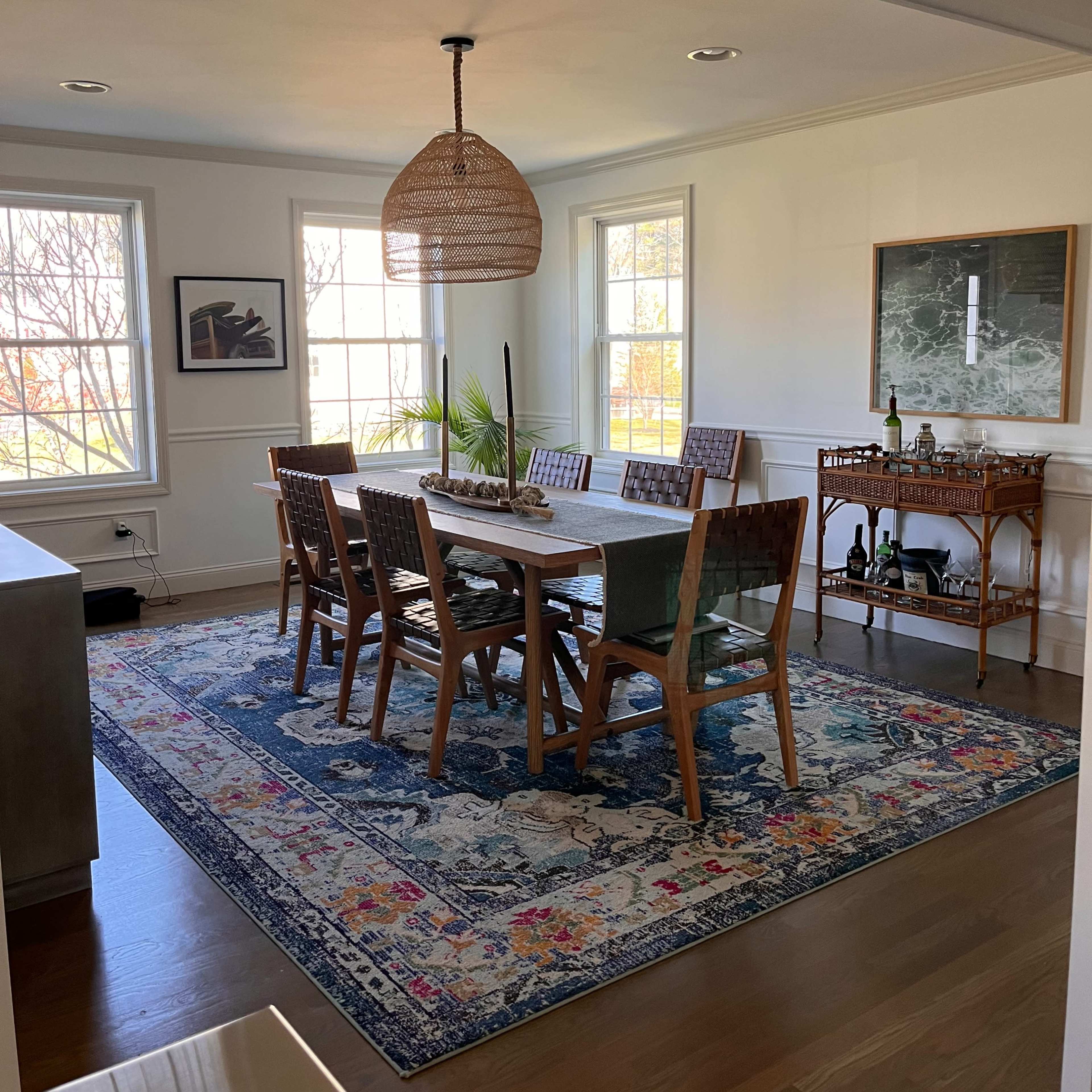A dining room features a long wooden table with eight chairs, a large area rug, and windows allowing natural light.