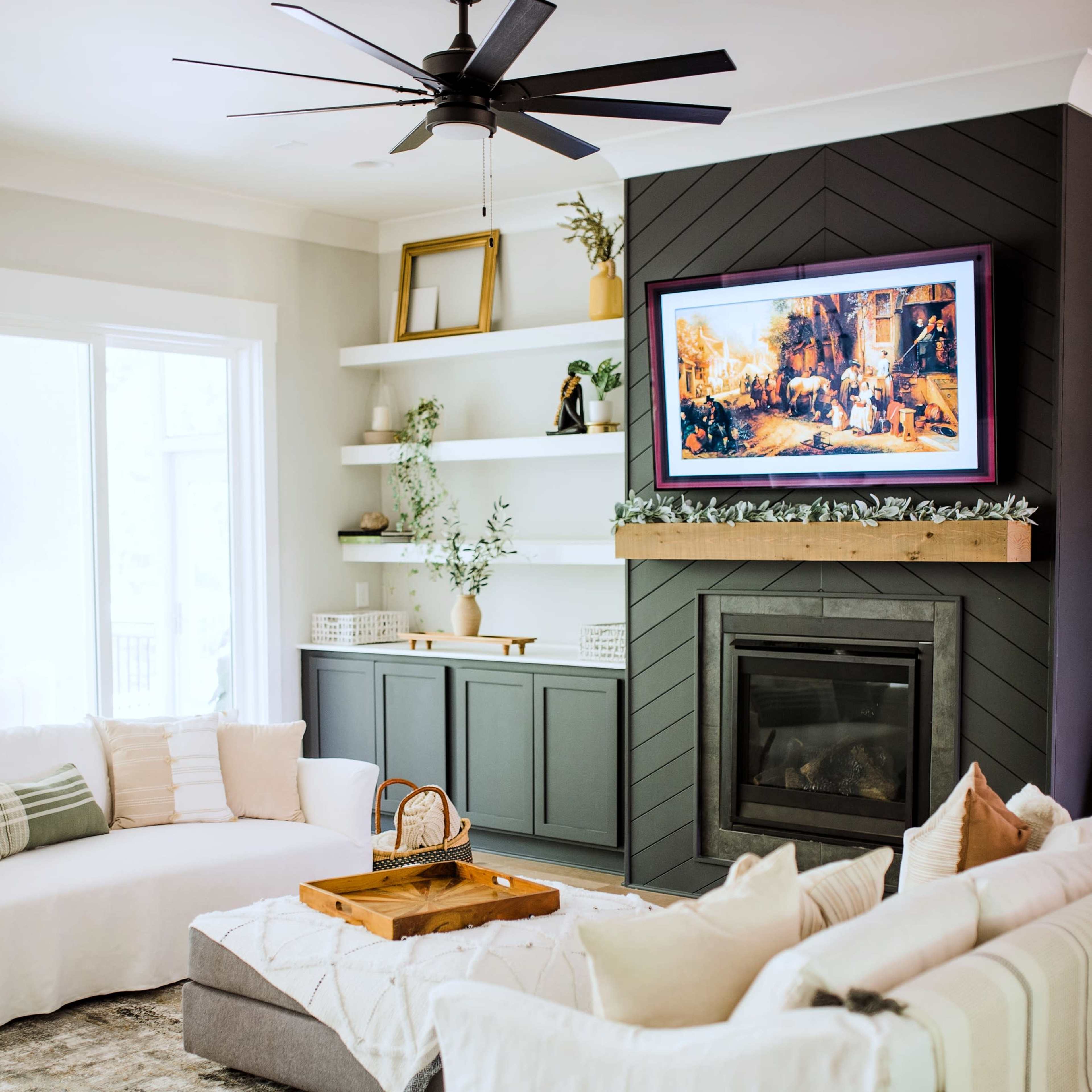 A modern living room features a ceiling fan, a fireplace with a television above it, and a sectional sofa arranged around a coffee table on a patterned rug.